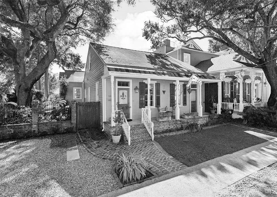 A black and white photo of a small house with a porch, steps, and a garden. The house has a gabled roof and a front door with a decorative fleur-de-lis. There are large trees on either side, and neighboring houses are visible in the background.