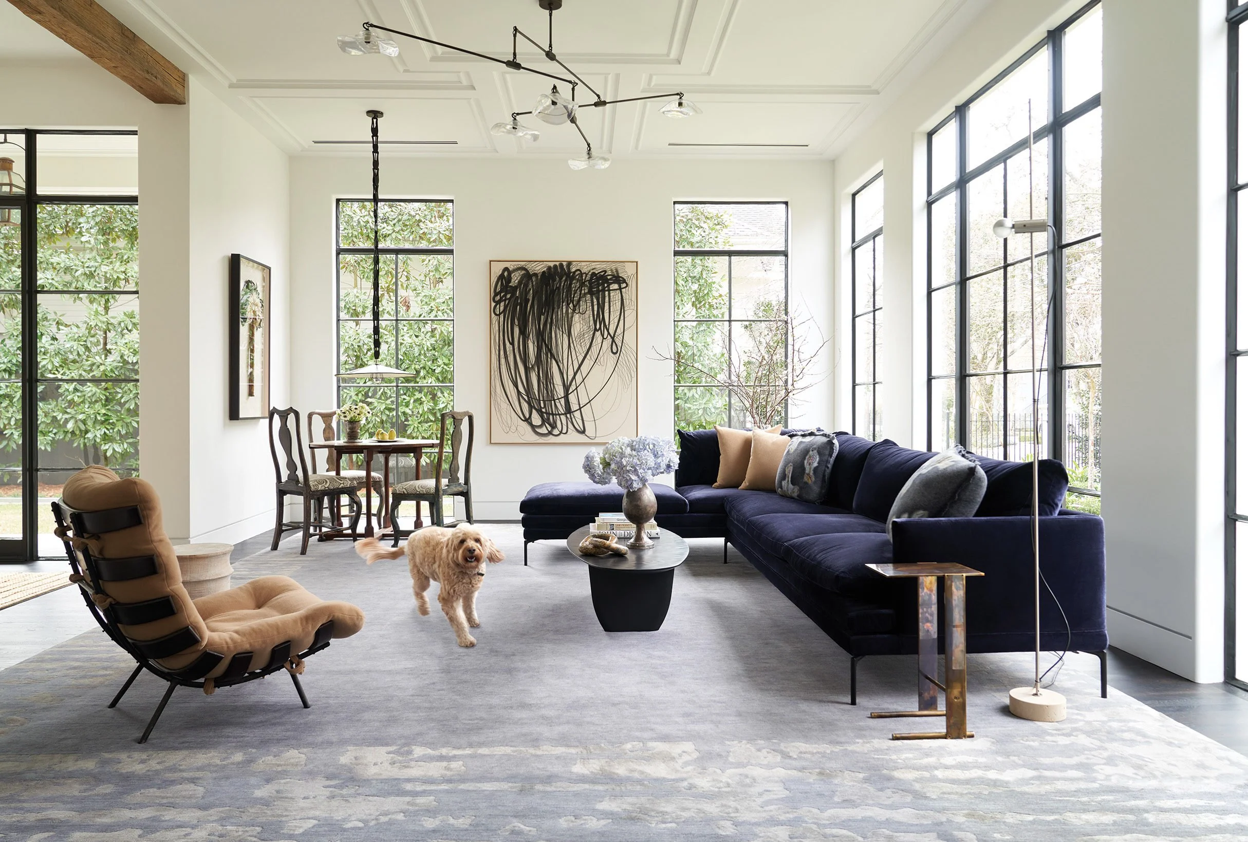 Modern living room with large windows, navy blue sectional sofa, beige armchair, coffee table with flowers, abstract artwork, dining area with chairs, and a dog on a gray rug.