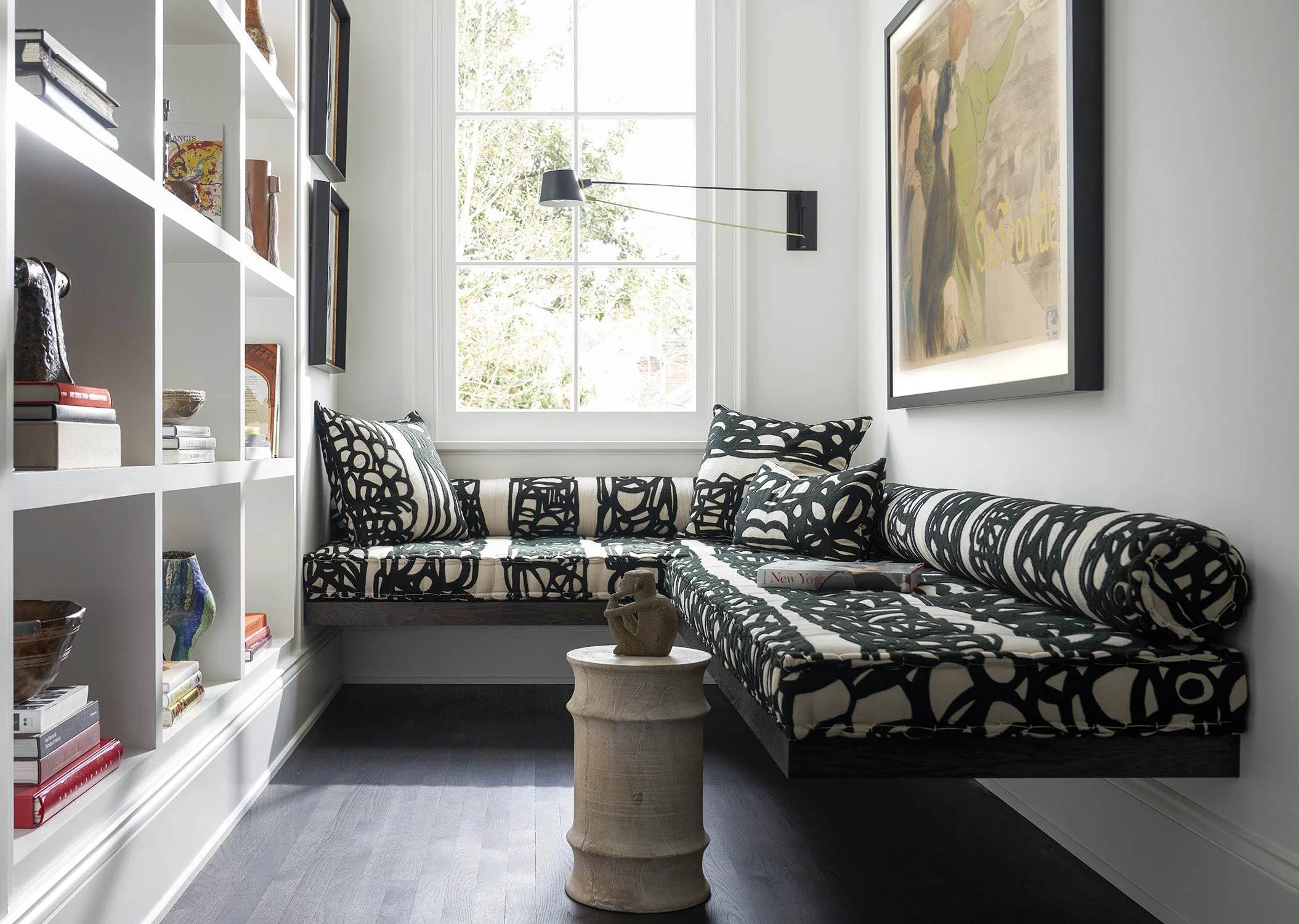 A cozy reading nook with a black and white patterned sectional sofa, decorative pillows, a small wooden side table with a sculpture, a large window allowing natural light, a book on the sofa, framed artwork on white walls, and a built-in white bookshelf filled with books and decorative items.
