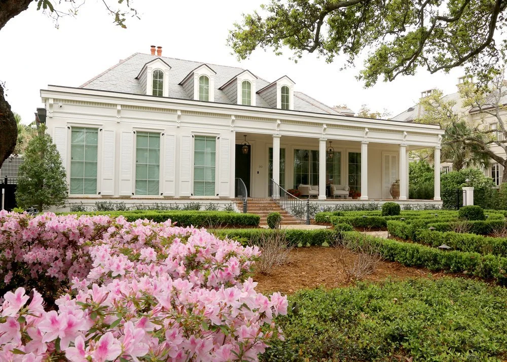 A large, white house with a gray roof, front porch with chairs and potted plants, surrounded by a garden with pink flowers, bushes, and trees.