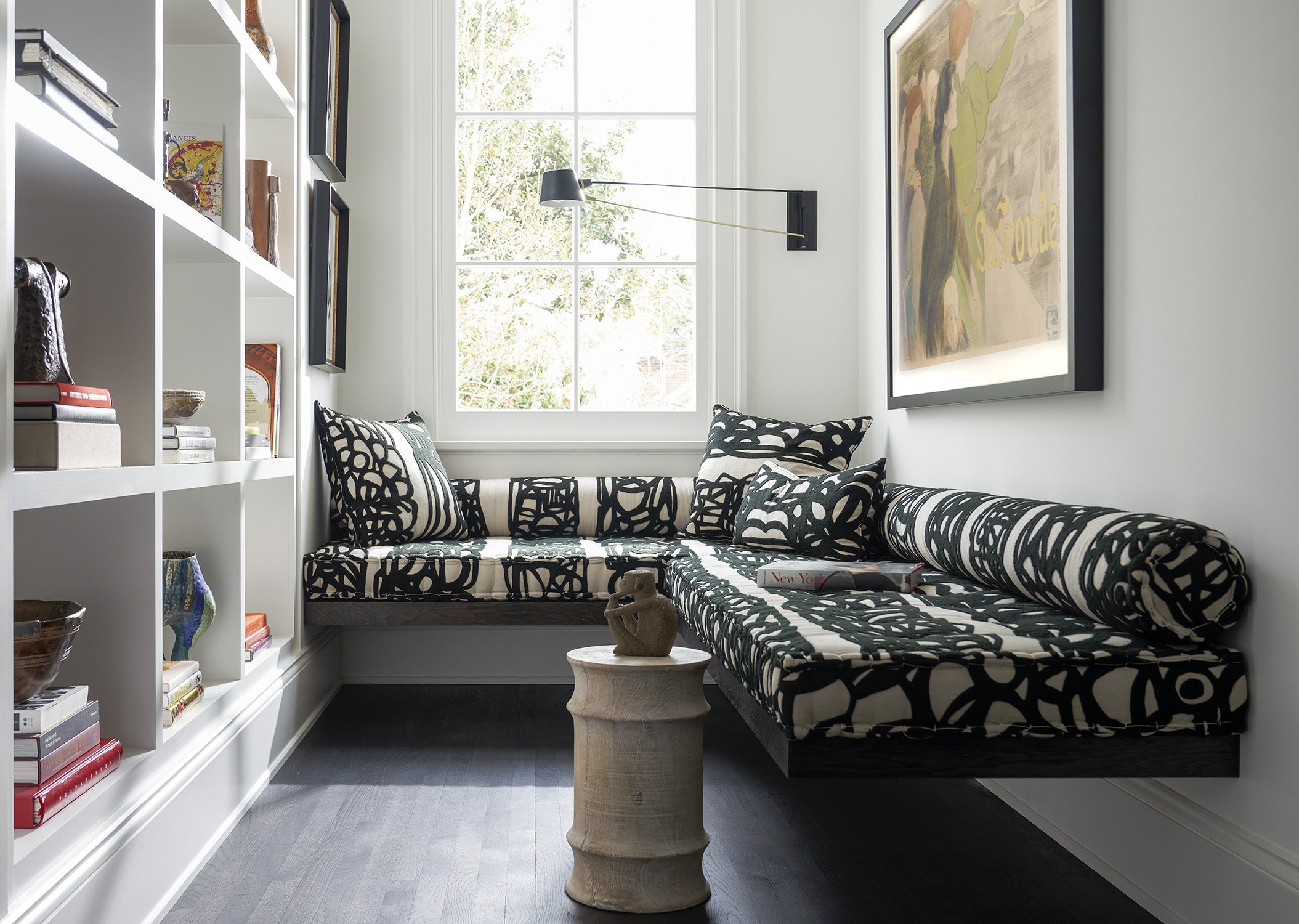 Small sitting area with black and white patterned cushioned window seat, a white bookshelf filled with books and decor, a small round side table with a sculpture, a large framed artwork on the wall, black wood flooring, and a window with natural ligh
