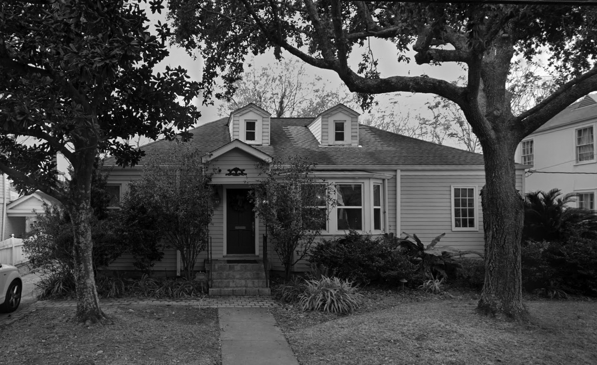Black and white photo of a house with a front pathway, stairs, two trees in the yard, and bushes surrounding the house.