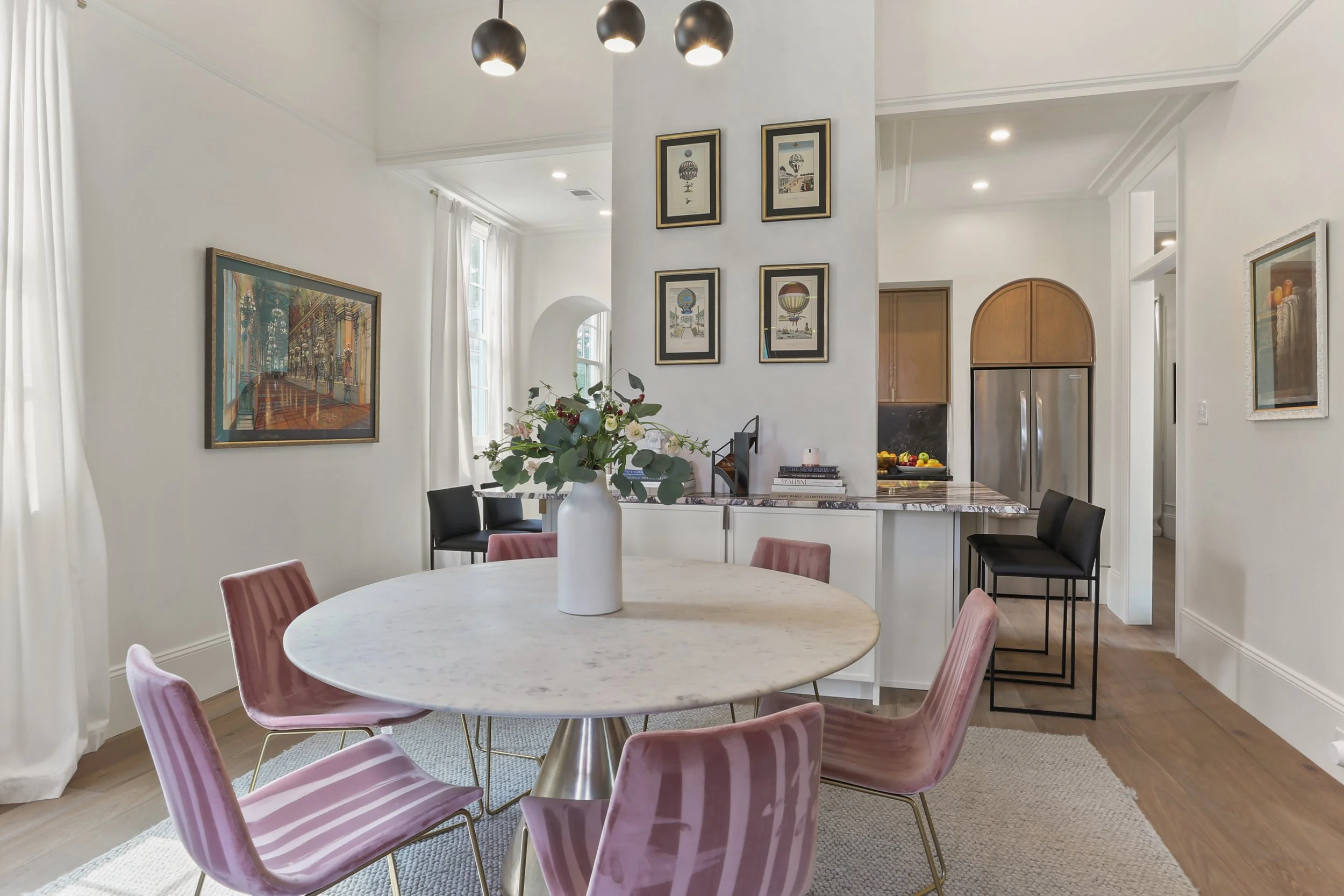 A dining area with a round marble table surrounded by six pink velvet chairs, a white vase with flowers in the center, and a wall with framed pictures and paintings, adjacent to a kitchen with black chairs and a marble countertop.