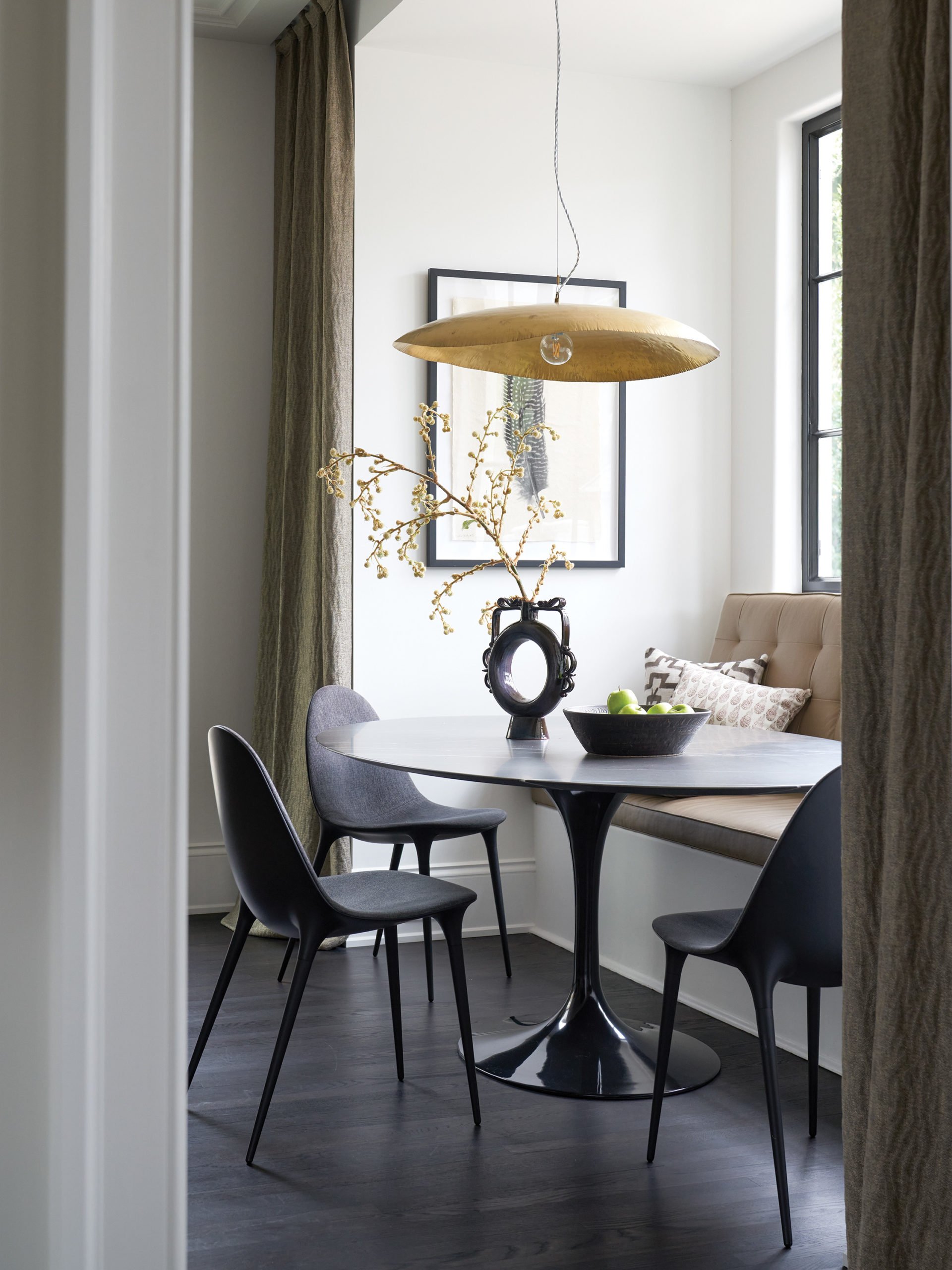A modern dining nook with a round table, three chairs, a built-in bench, and a framed art piece on the wall. A gold pendant light hangs above the table, and a black vase with a branch and berries decorates the table. Large windows with curtains provi