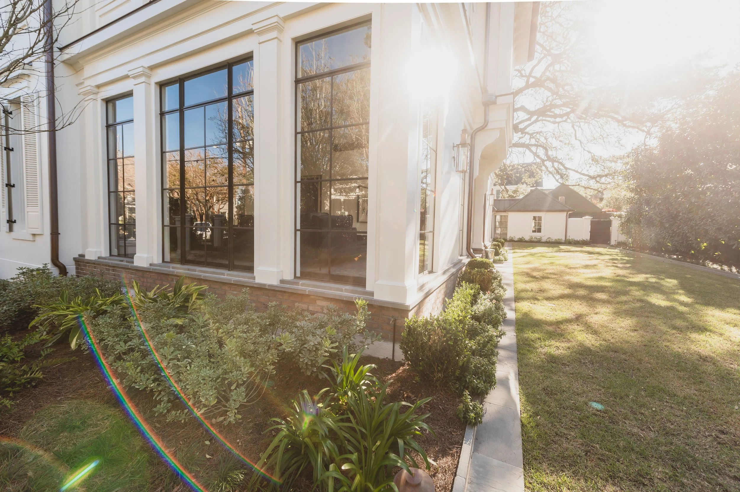 Sunlight shining over a landscaped backyard with a well-maintained lawn and bushes, next to a white house with large windows.
