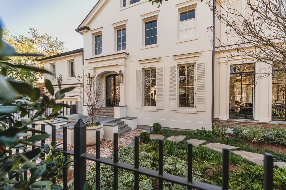White house with multiple windows, shutters, and a front garden enclosed by a black fence.