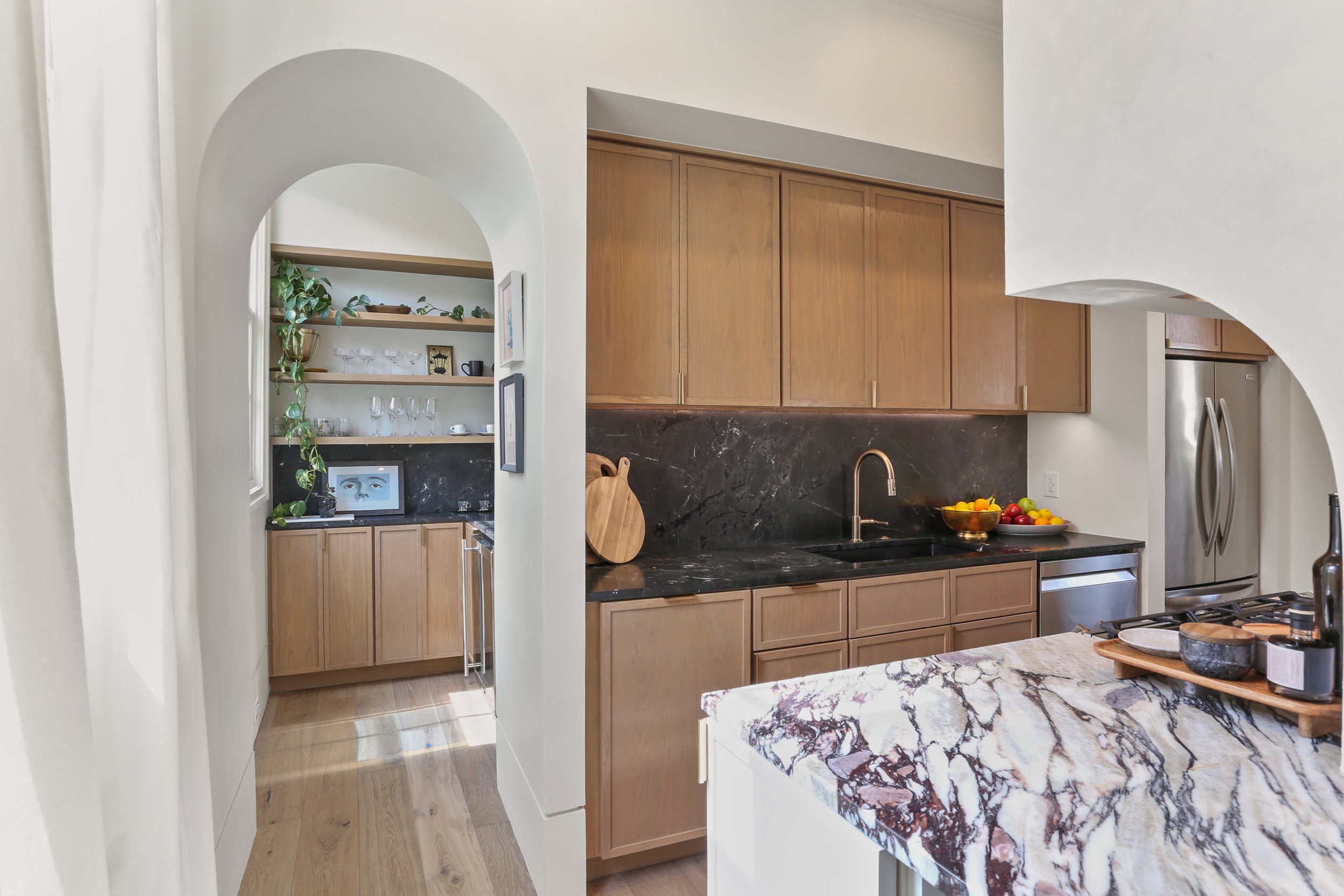 Kitchen with wooden cabinets, black marble countertops, fruit bowl, and a stainless steel refrigerator, with a view into a small but stylish nook with open shelves and decorative items.