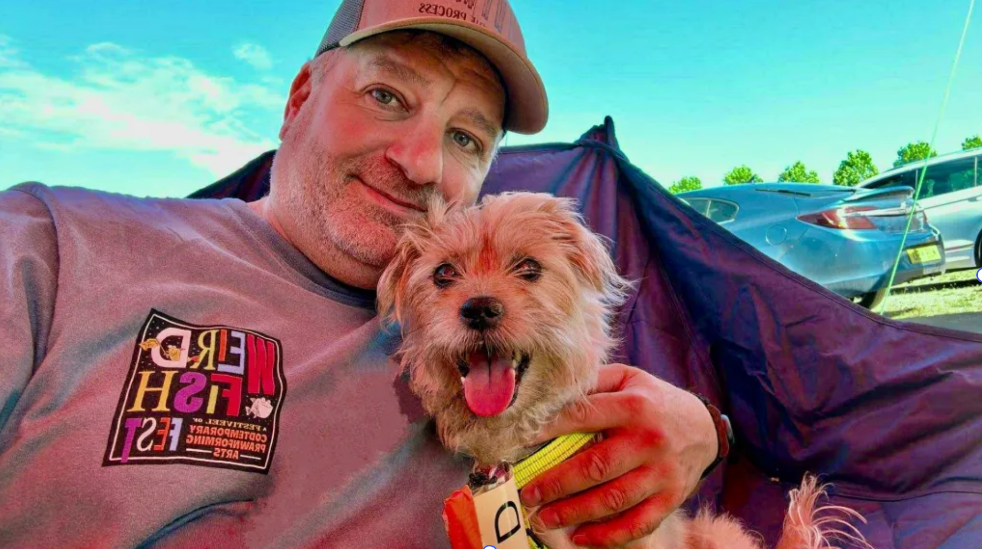 A man smiling and holding a smiling small dog with a yellow collar. They are outdoors with parked cars and a camping tent in the background.