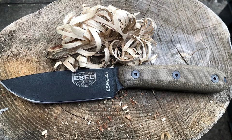 A black fixed-blade knife with a tan handle lying on a wooden surface next to wood shavings.