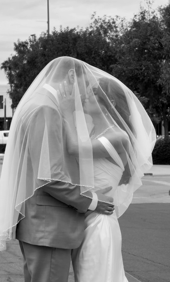 A bride and groom kiss under a veil in a black and white wedding photo outdoors.