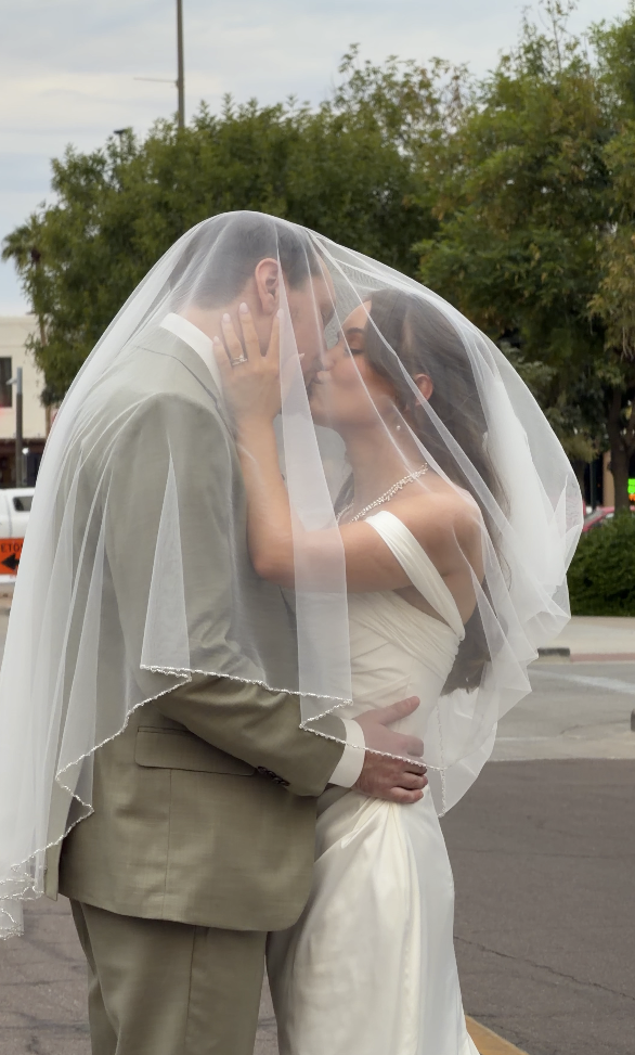 Bride and groom share a kiss under a veil outdoors with trees and a street in the background.