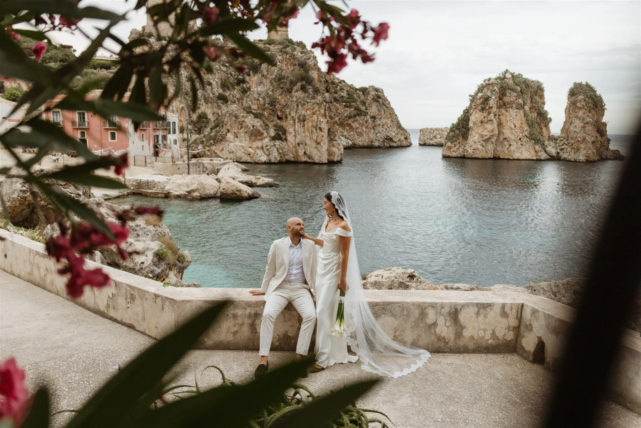 A bride and groom are posing by a waterfront with rocky cliffs and small islands in the background during their wedding photoshoot.