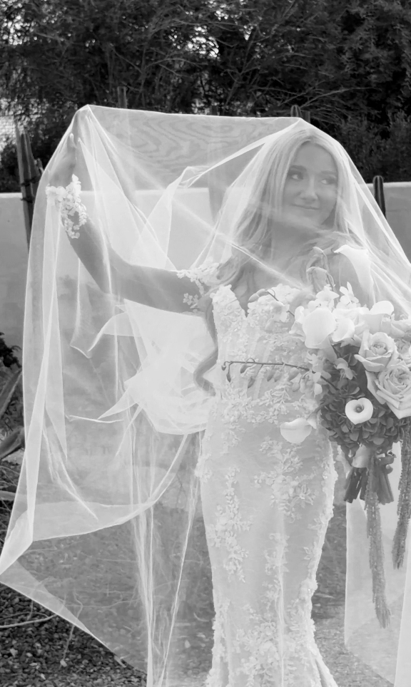 Brunette bride in a lace wedding dress holding a bouquet of flowers, standing outdoors with a veil covering part of her face and hair.