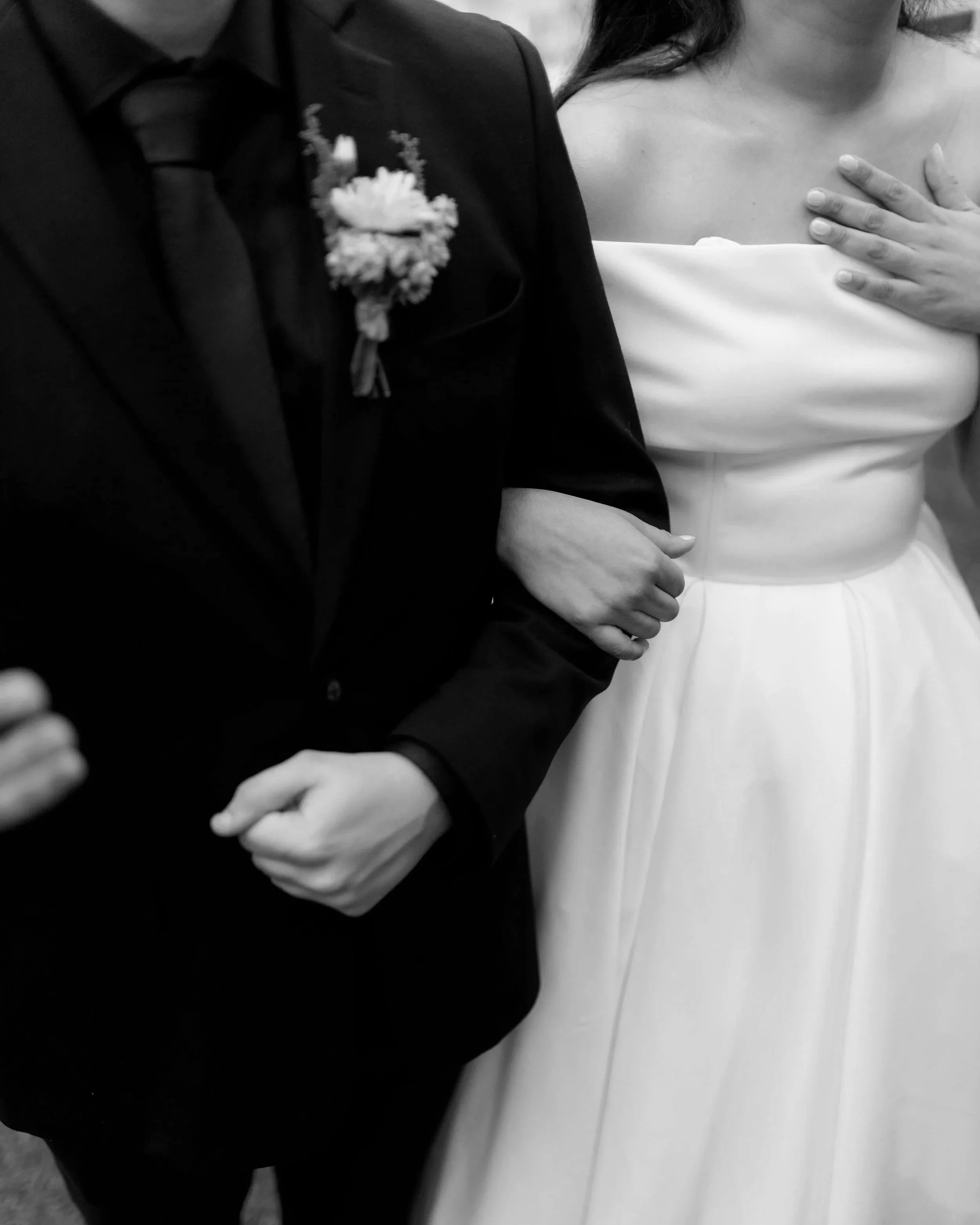 A black and white photo of a bride and groom at their wedding, with the groom in a dark suit and the bride in a strapless white dress, holding each other's arms.