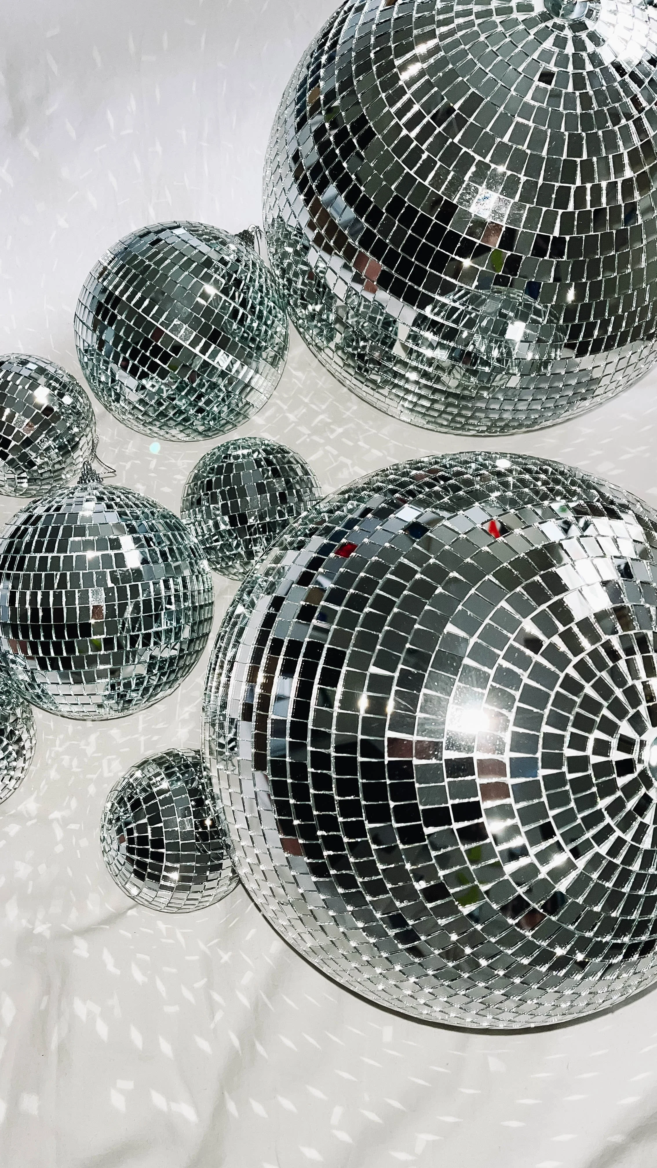 Multiple disco balls of various sizes hanging against a white background, reflecting light and casting shimmering reflections.