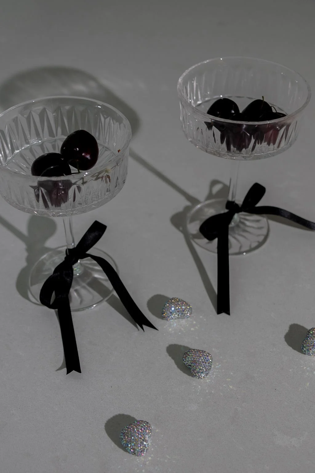 Two crystal champagne glasses with black ribbon bows, filled with cherries, on a white surface. Glittery heart-shaped decorations are scattered around, and there are shadows cast by the glasses.
