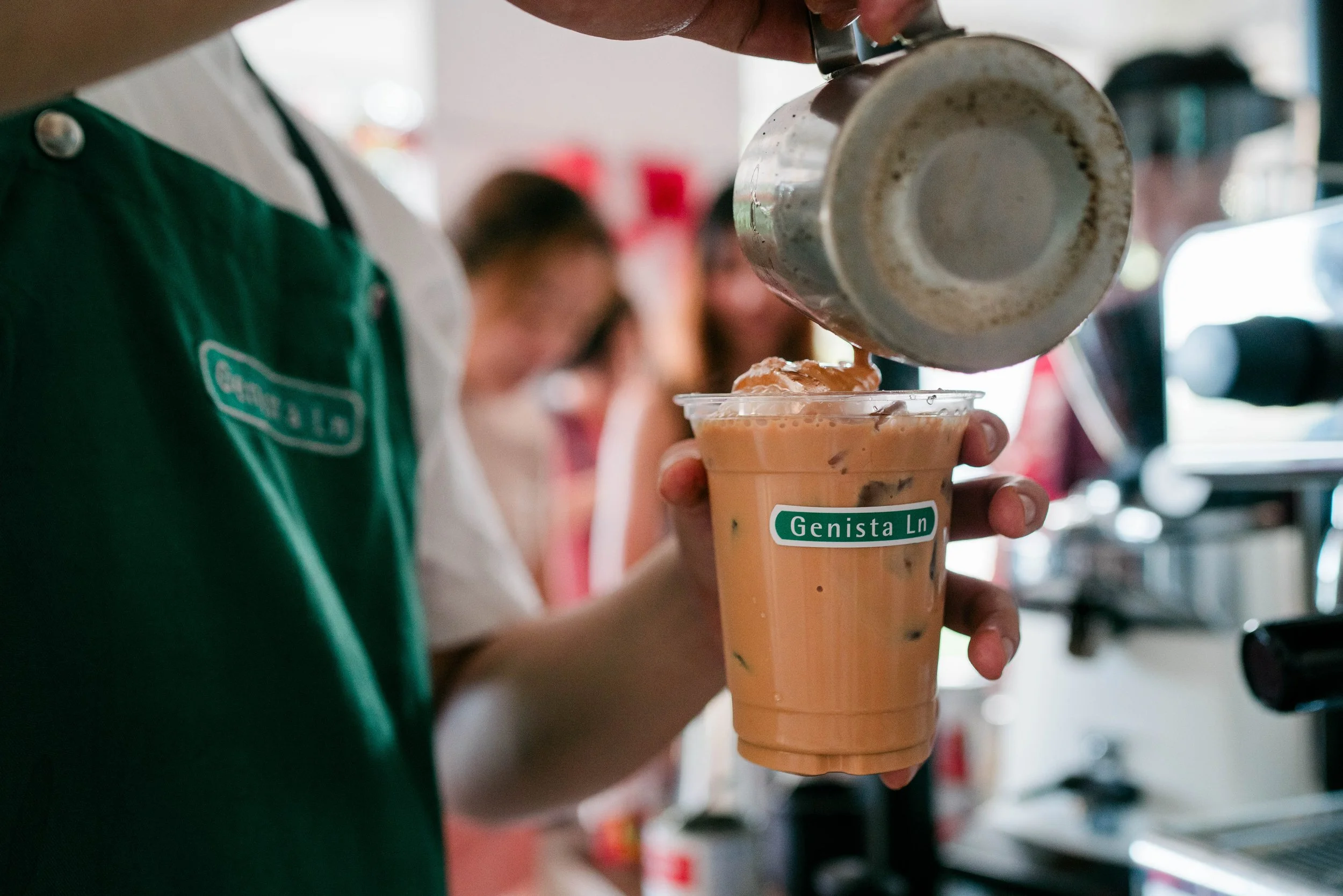 Barista pouring ice into a plastic cup of iced coffee or tea at a cafe, with other customers in the background.