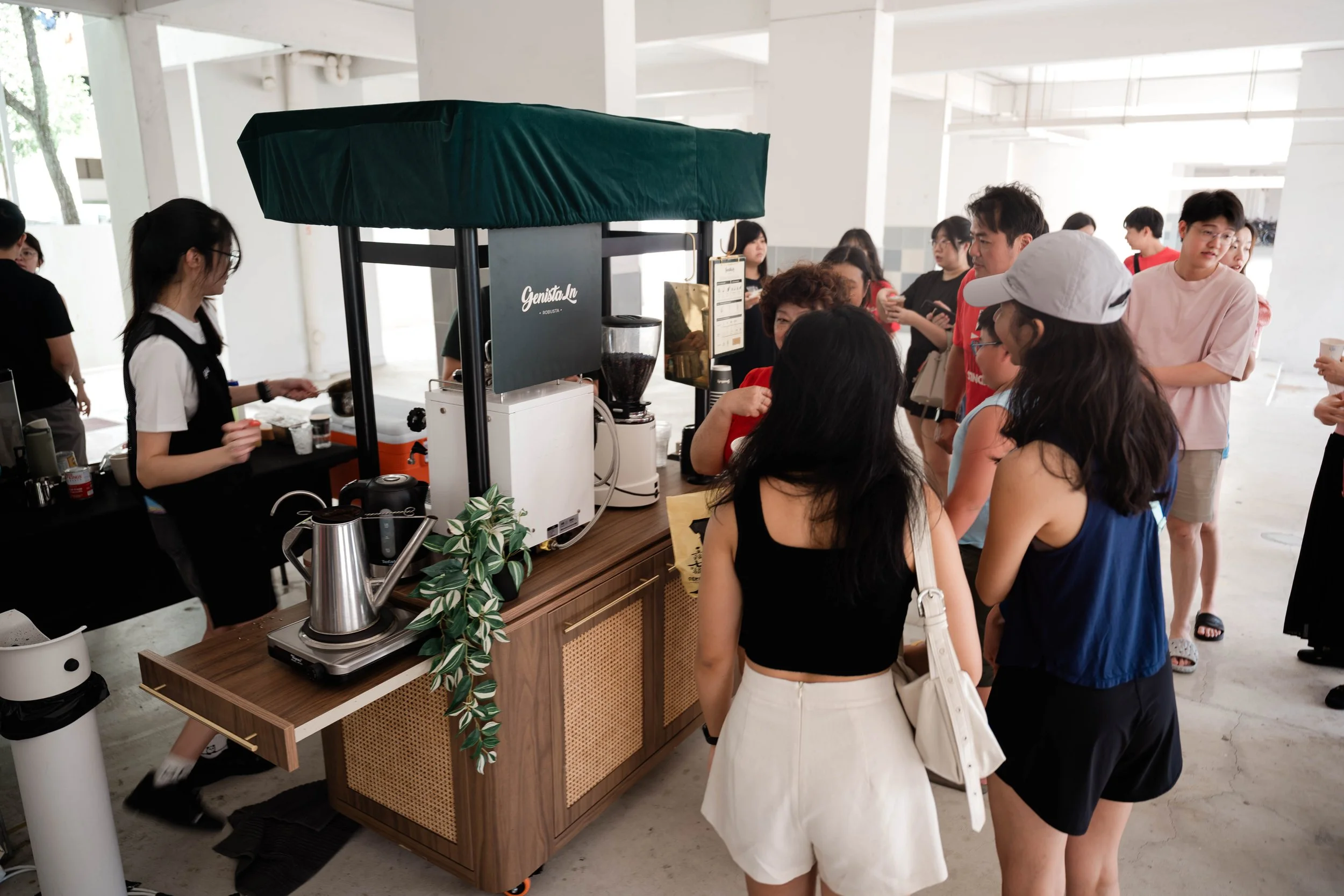 People standing in line at a coffee stand under a void deck, with barista preparing drinks and coffee equipment on the counter.