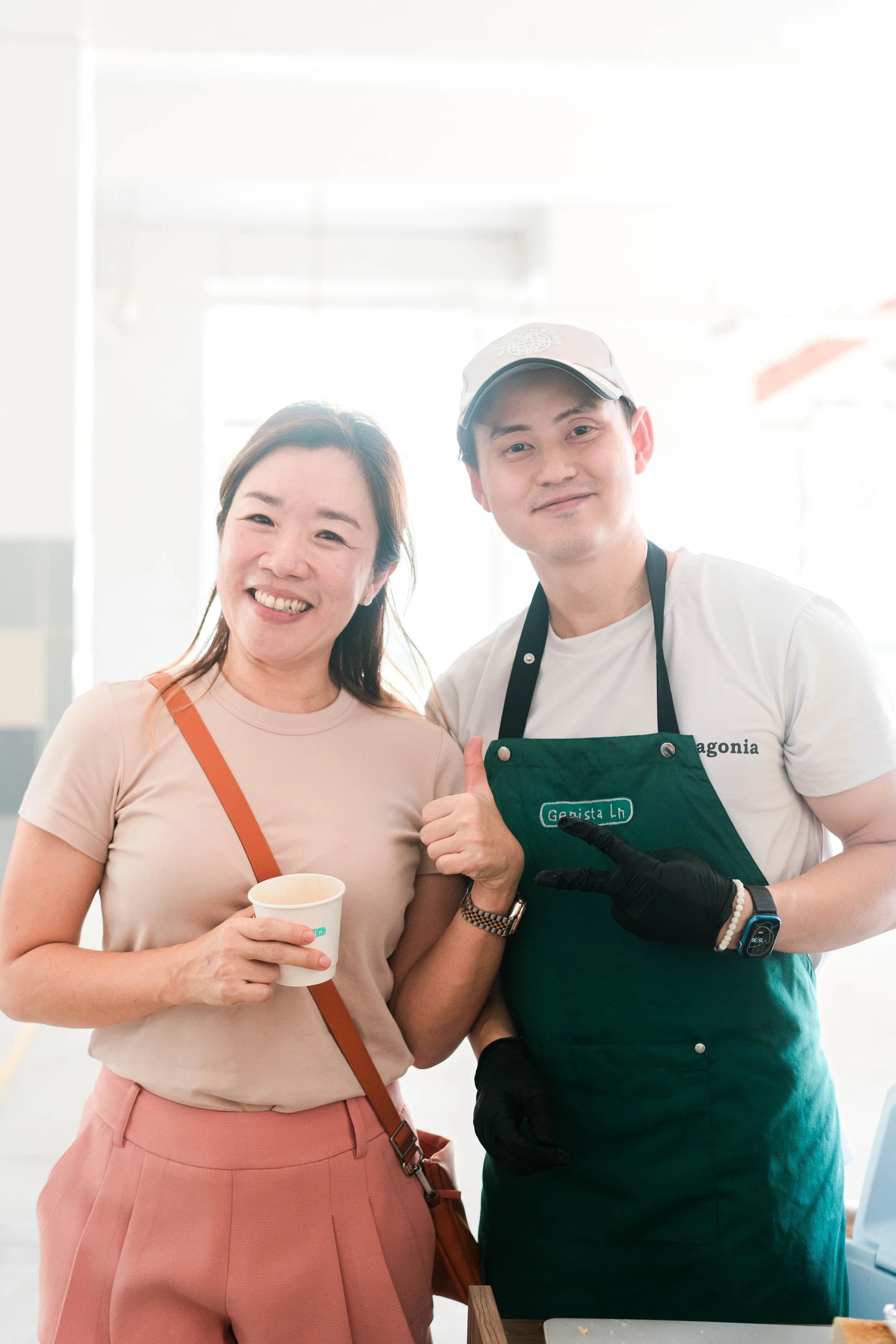 A smiling woman holding a paper cup standing next to a man in a green apron making a peace sign in a bright indoor setting.