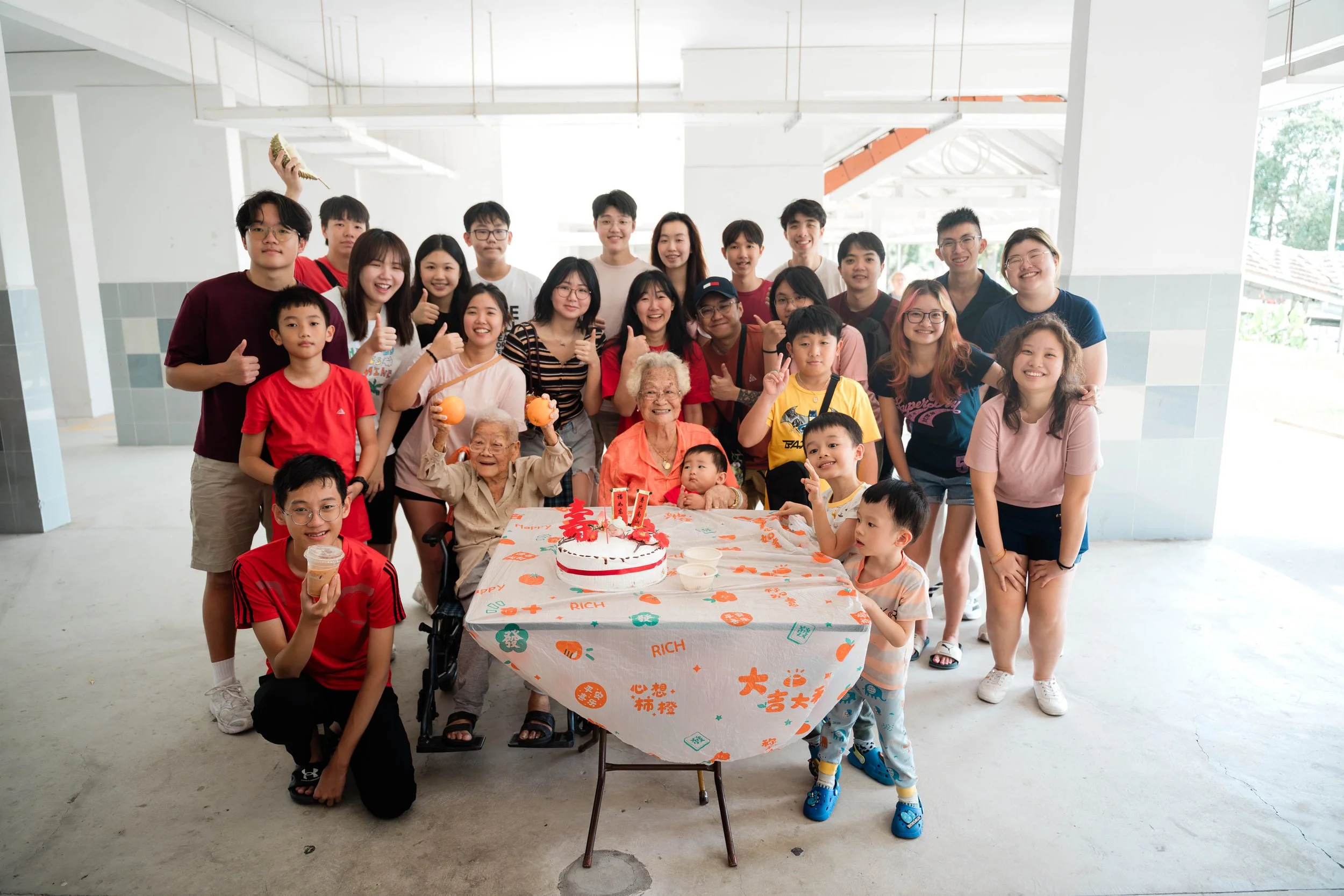 Group of people gathered around a table with a birthday cake, celebrating a birthday with elderly women, children, and adults in a bright, open space.