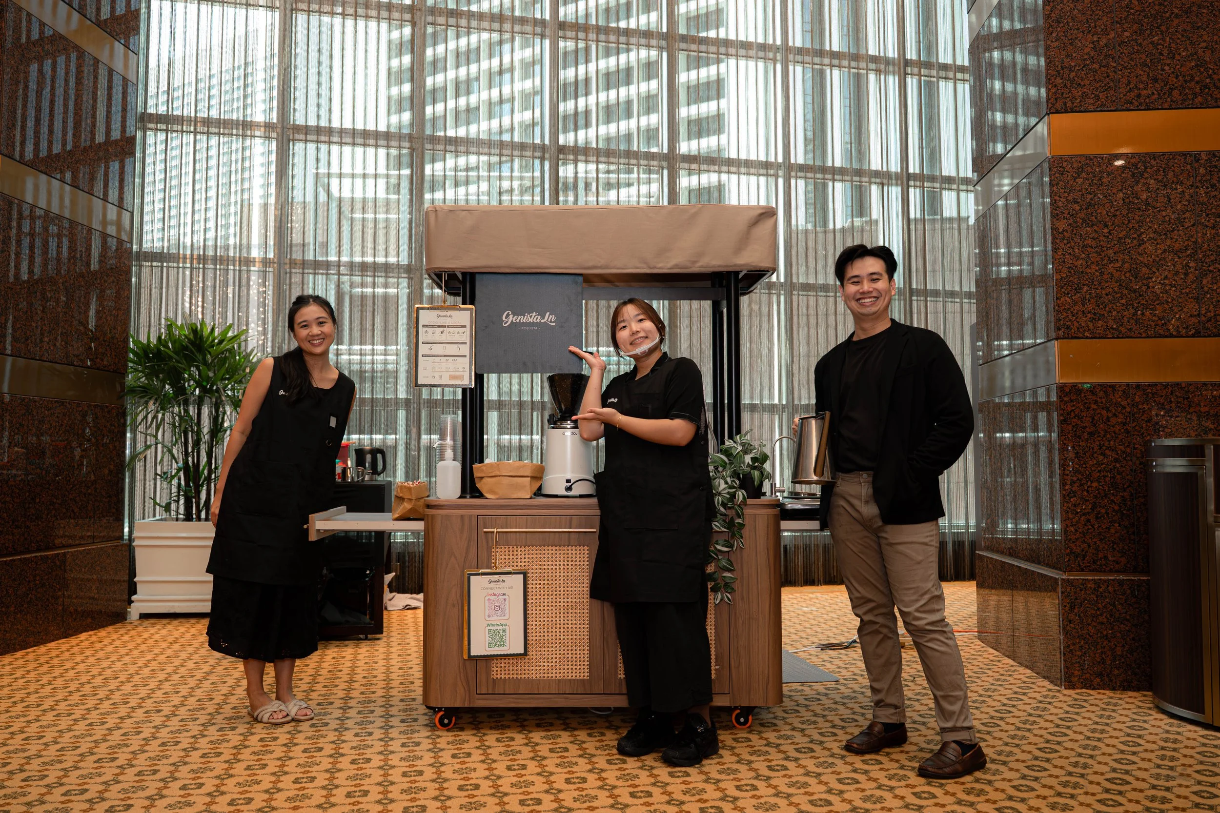 Three people standing behind a coffee stand inside a building with large windows. The two women and one man are smiling. The coffee stand has a coffee grinder, cups, and other items. The background shows tall buildings outside.