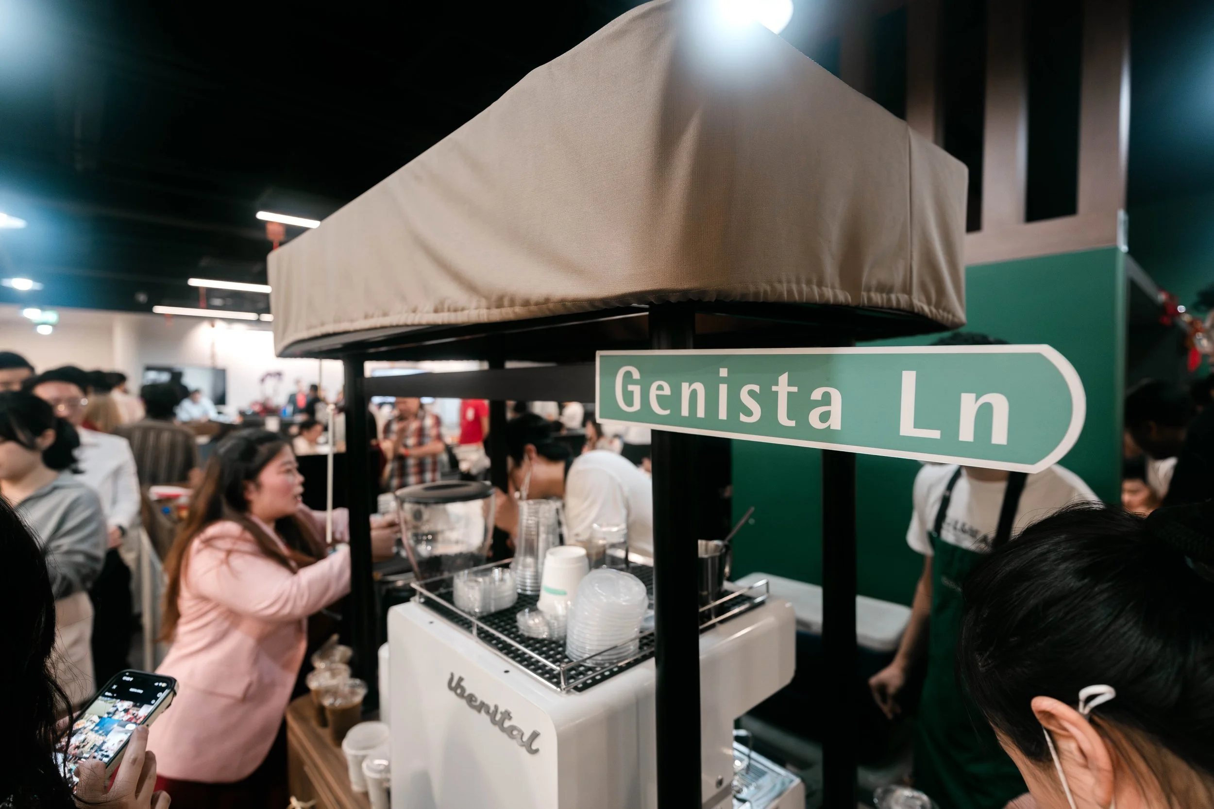 Crowd of people at a Kopi Cart with a sign reading 'Genista Ln' in an indoor setting.