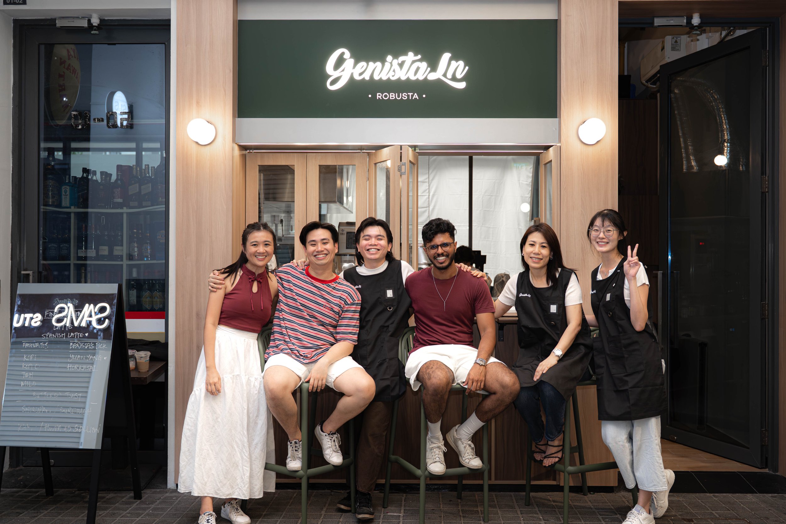 Group of six people smiling and posing together in front of a restaurant named Genista Ln Robusta.