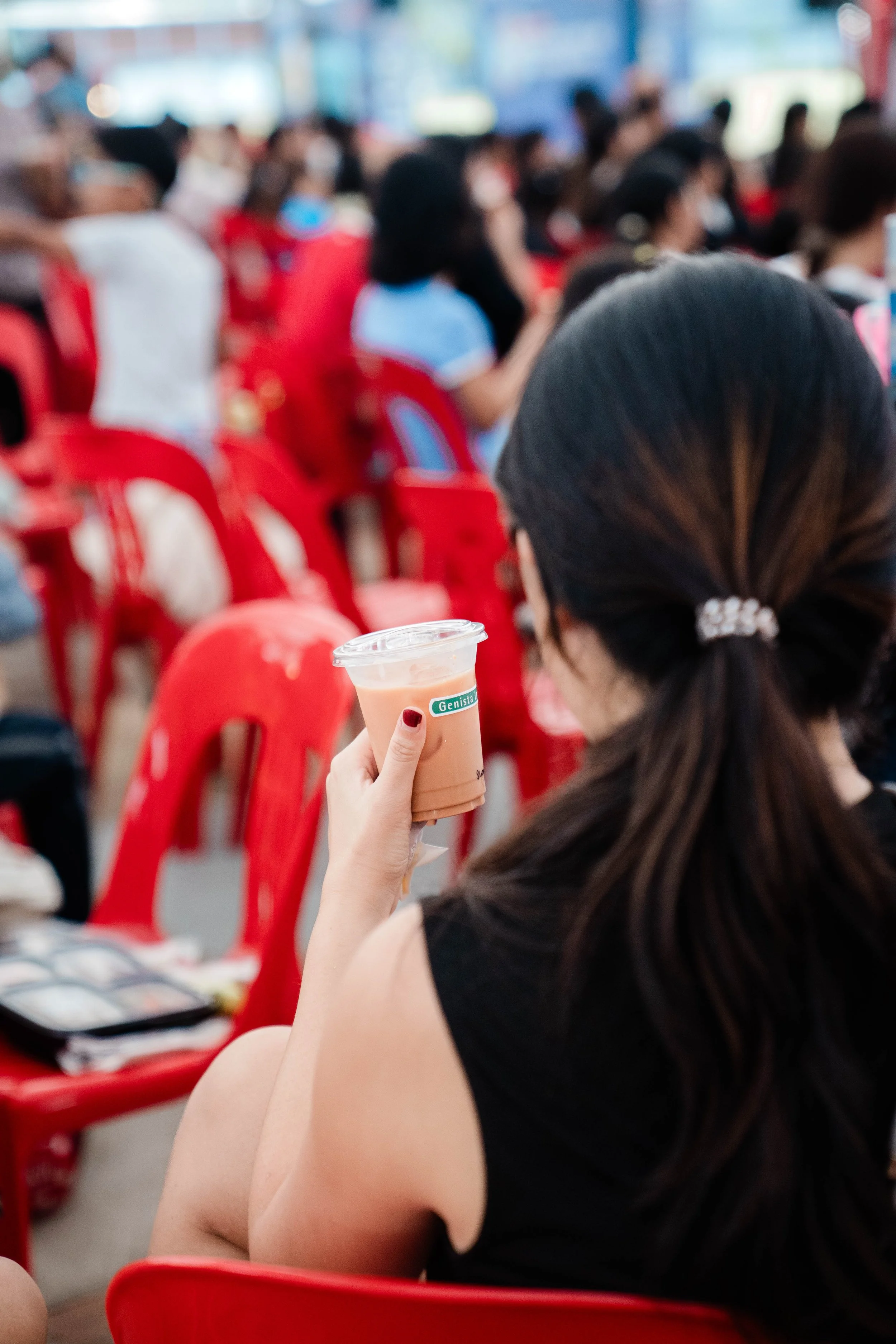 A woman with long dark hair tied back with a hair clip, wearing a black sleeveless top, is sitting on a red plastic chair in a crowded outdoor setting. She holds a cup of iced beverage with a straw, and several people are sitting on similar chairs behind her.