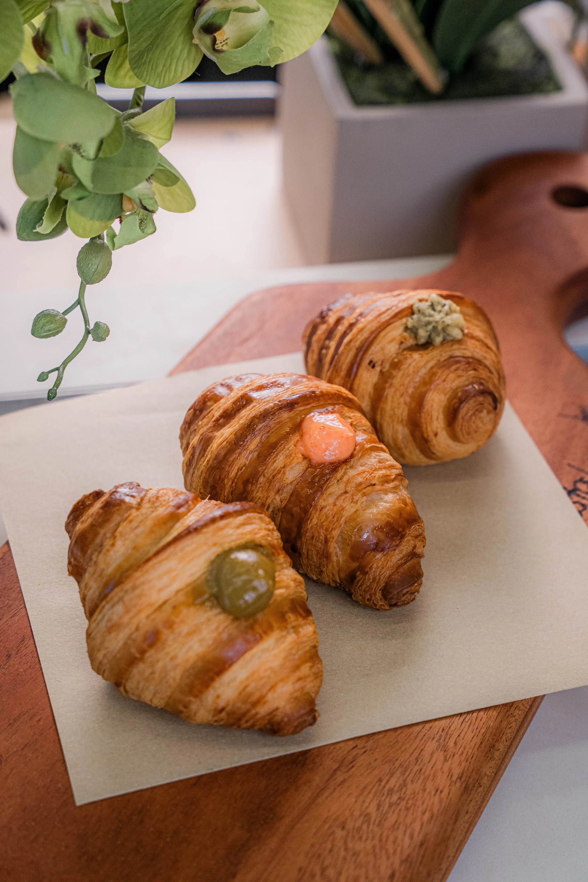 Three croissants on a piece of parchment paper on a wooden board, each topped with different colored dollops of sauce or preserve, with a green plant above in a white pot in the background.