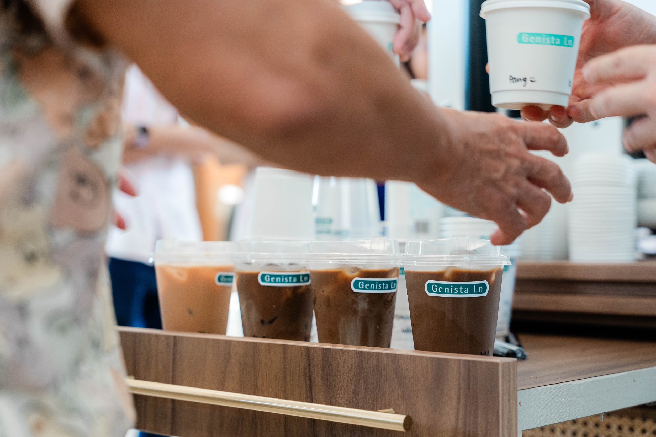 People serving iced coffee drinks in plastic cups with labeled stickers that say "Genista Ln", on a wooden tray at a coffee stand.