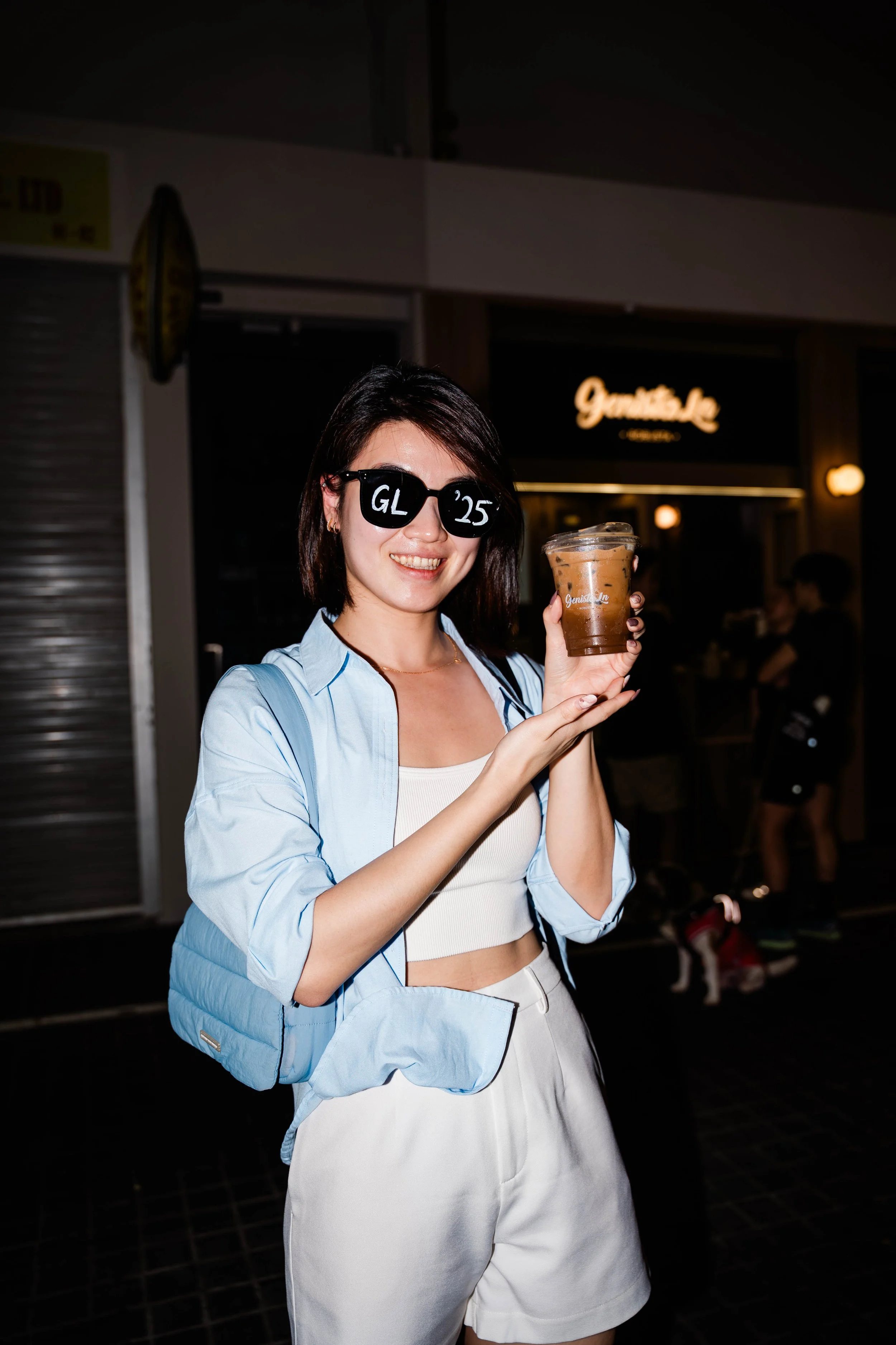 A young woman smiling, wearing sunglasses with 'GL 25' on the lenses, holding a cup of iced coffee in front of a dark background with a sign that reads 'Gourmet Inn' at night.
