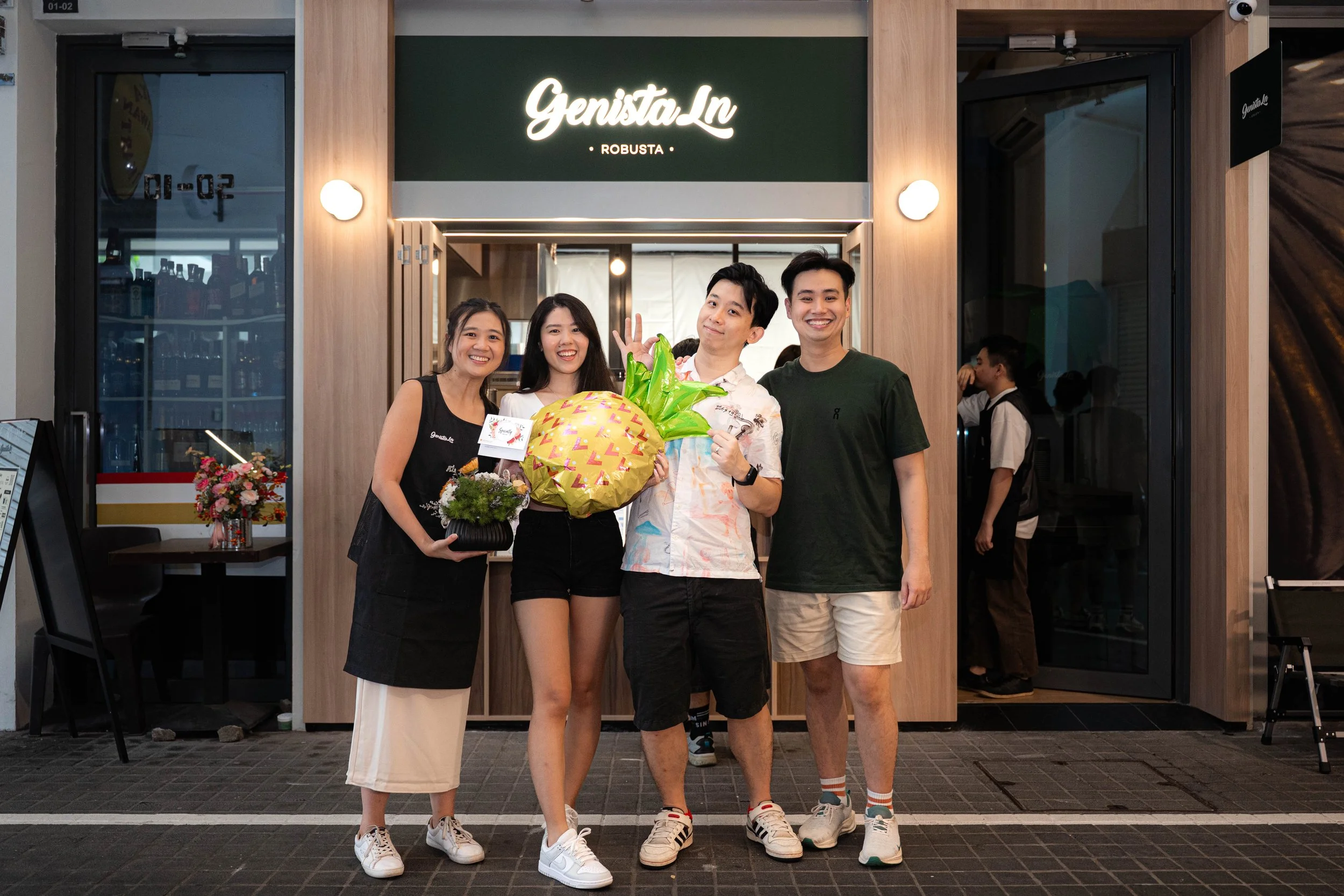 Group of four young adults standing outside a restaurant entrance, celebrating with flowers and a gift, smiling and waving at the camera.