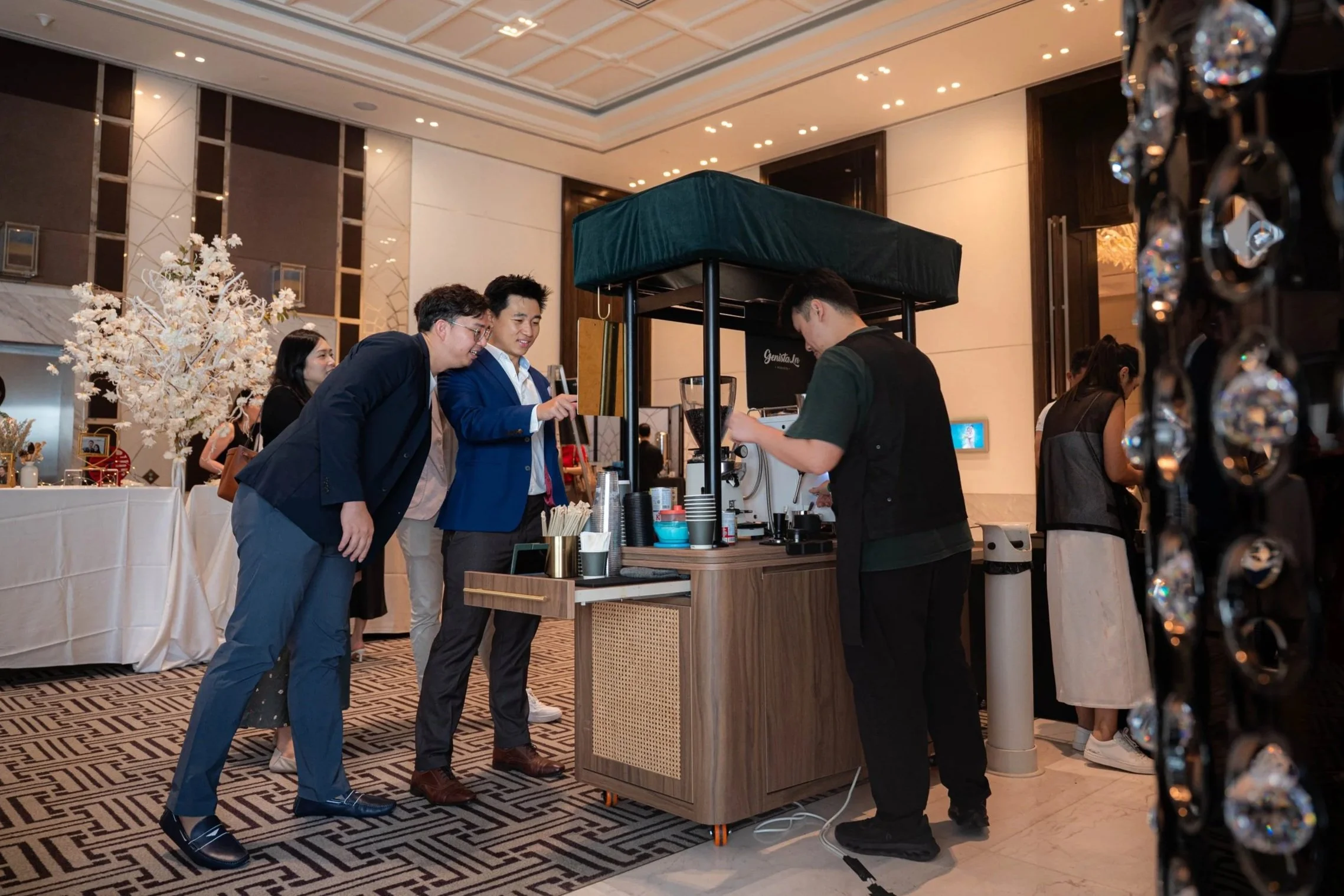 Group of people ordering coffee at Genista Ln Kopi Cart in a hotel lobby in Westin, with ornate decor and floral arrangements visible in the background.