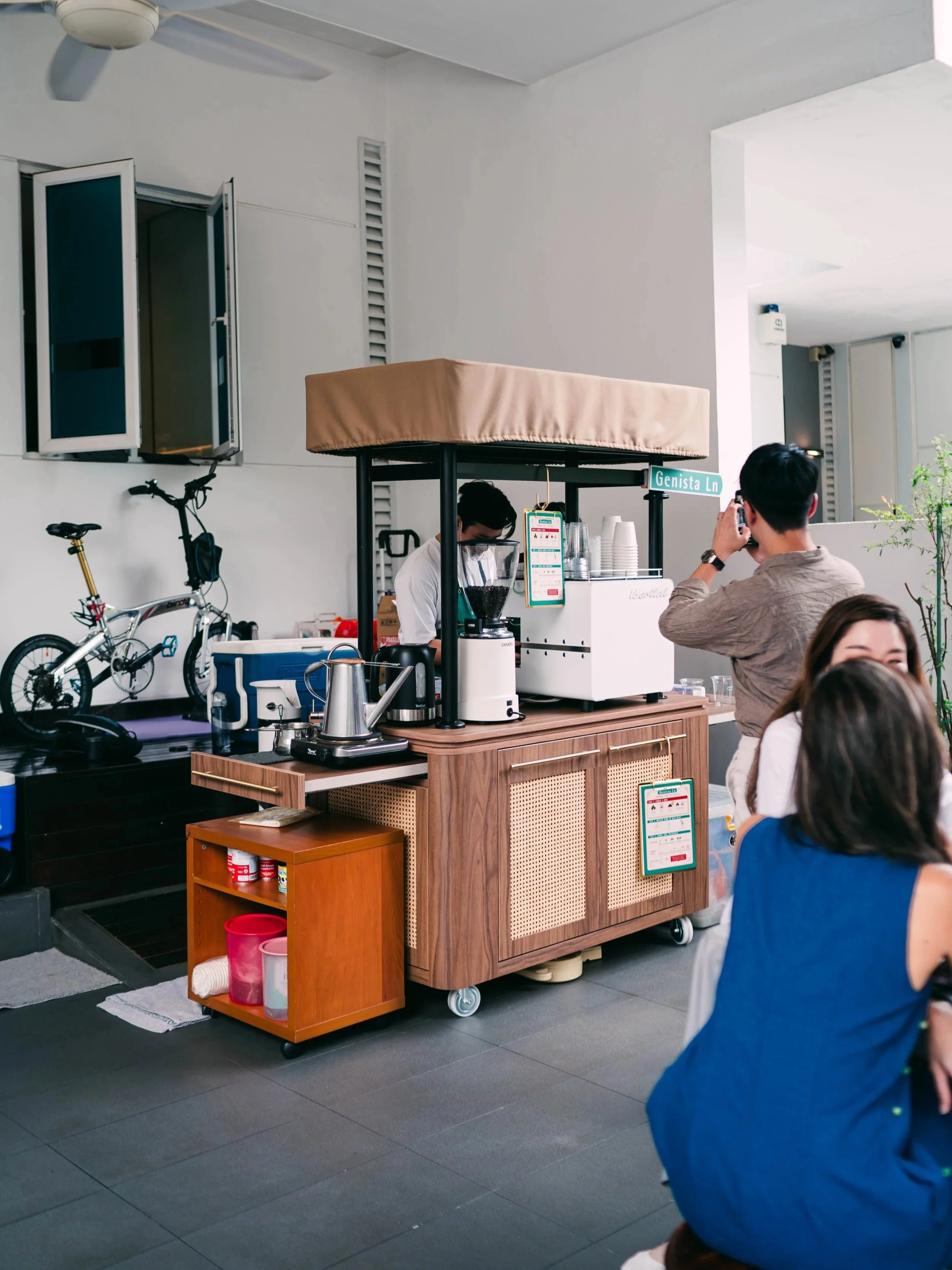 Genista Ln Kopi Cart with a barista preparing coffee, at a community event with a birthday party at a landed property house. There are customers ordering, a bicycle and some supplies nearby.