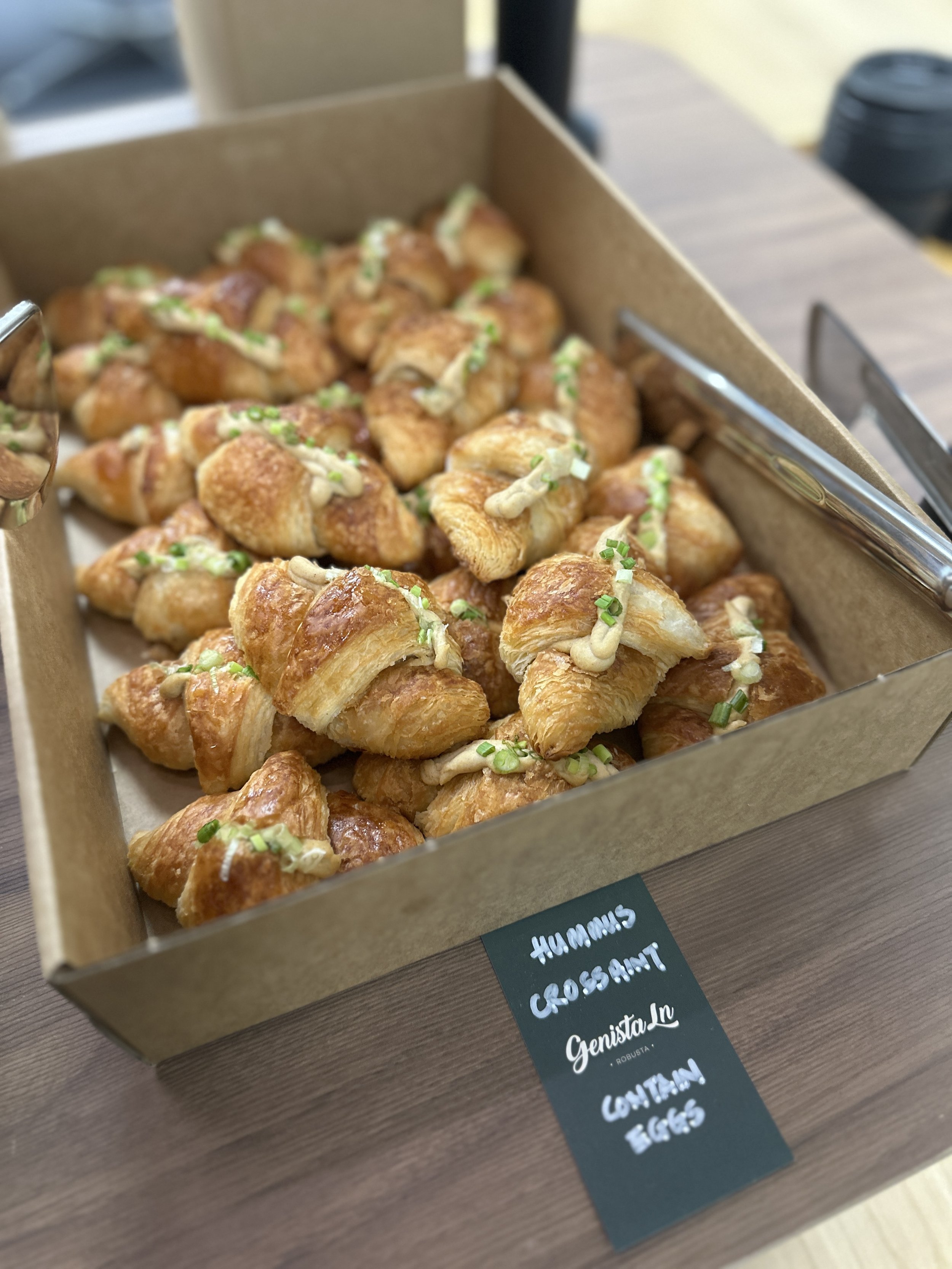 A tray of small, golden-brown croissant-shaped pastries topped with piping of sauce and chopped green onions, labeled as 'Hummin Crossaint' contained eggs.