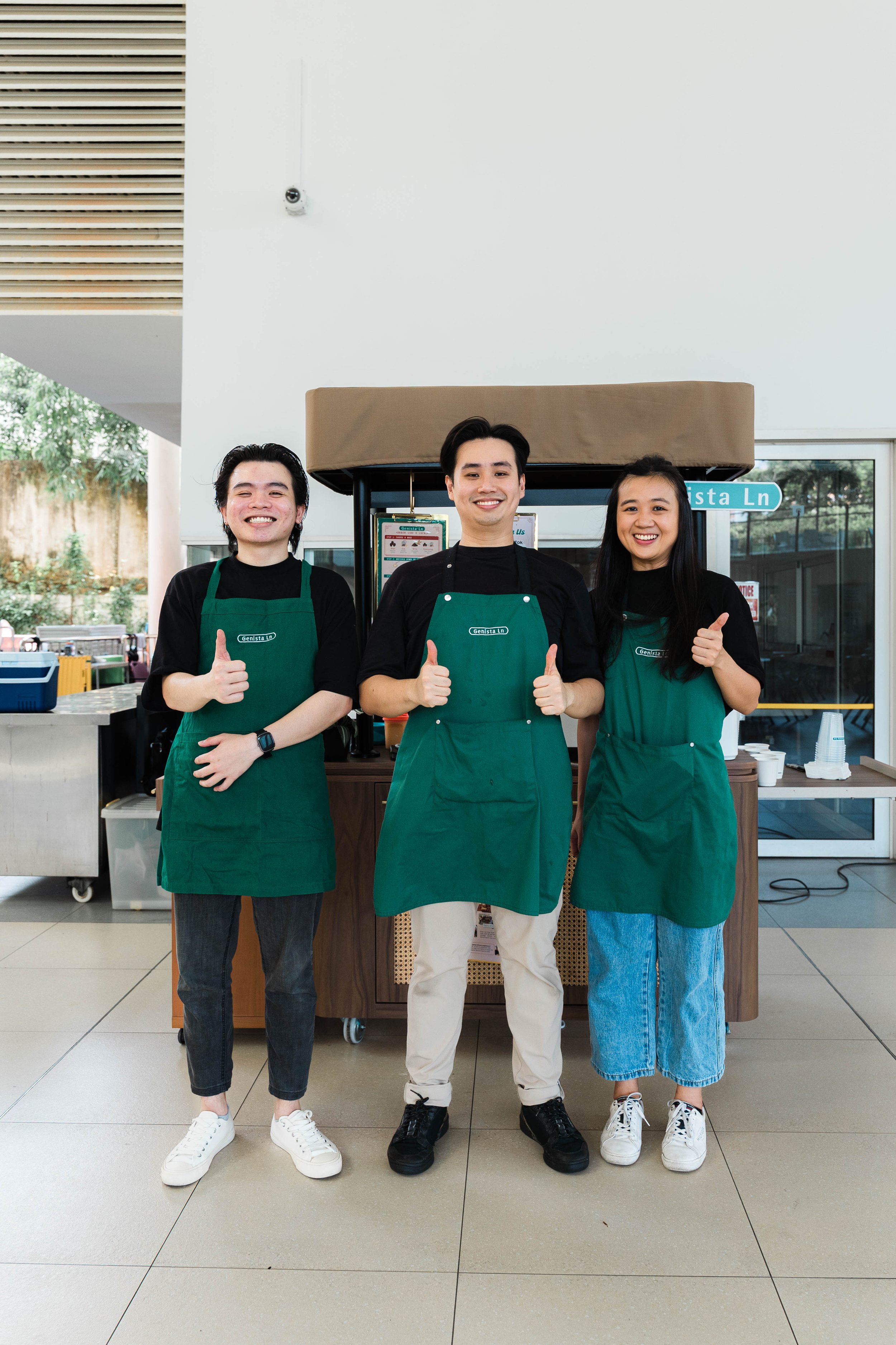 Three smiling employees wearing green aprons standing behind a cafe counter, showing thumbs up.