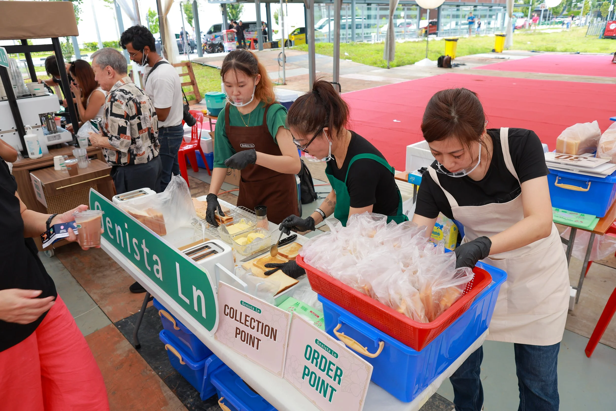 Three women working at a food stall with signs labeled 'Collection Point' and 'Order Point', serving customers at an event, with people in the background.