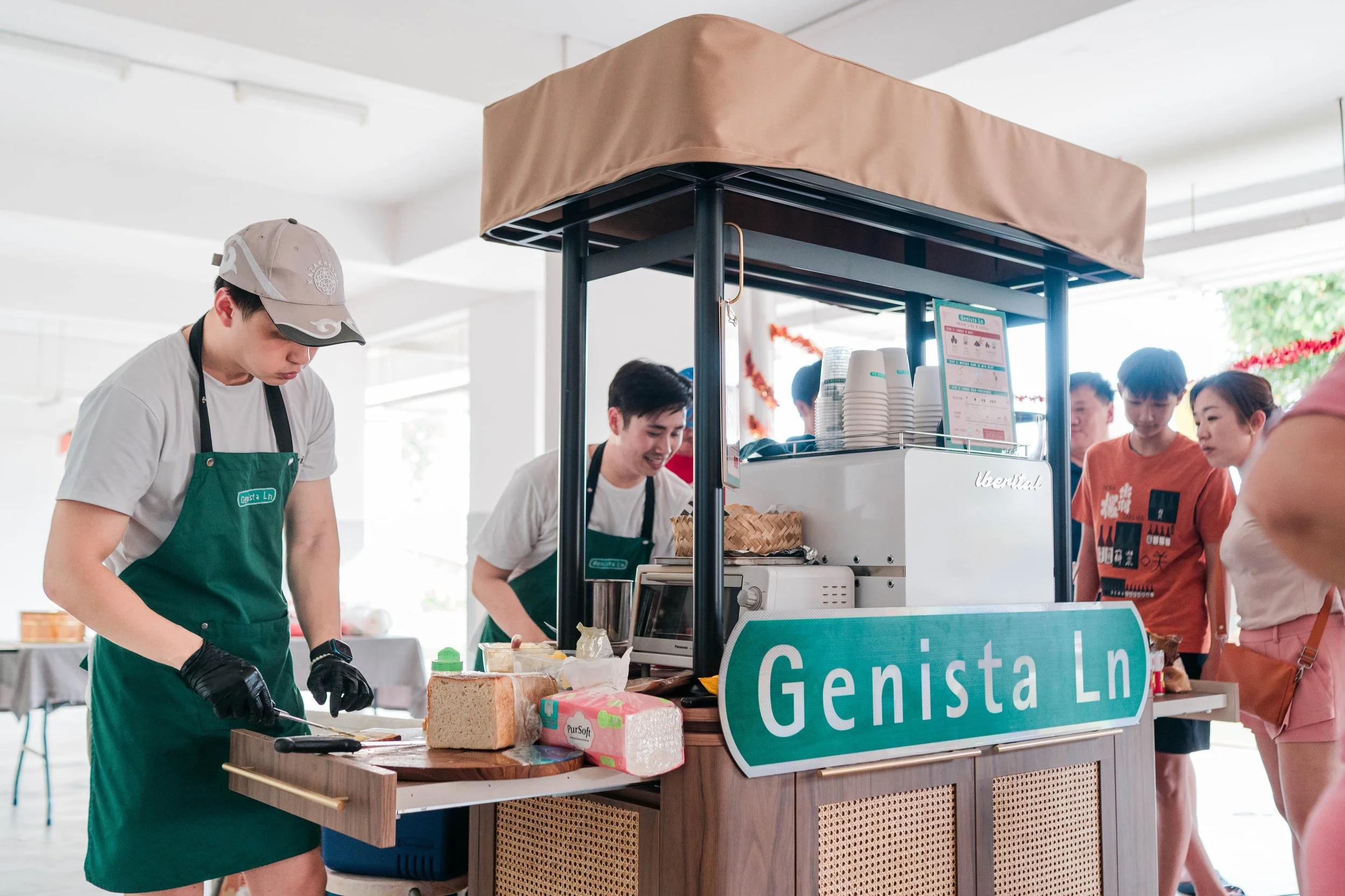 People preparing food at a street food stall with a sign that reads 'Genista Ln' in an indoor setting.
