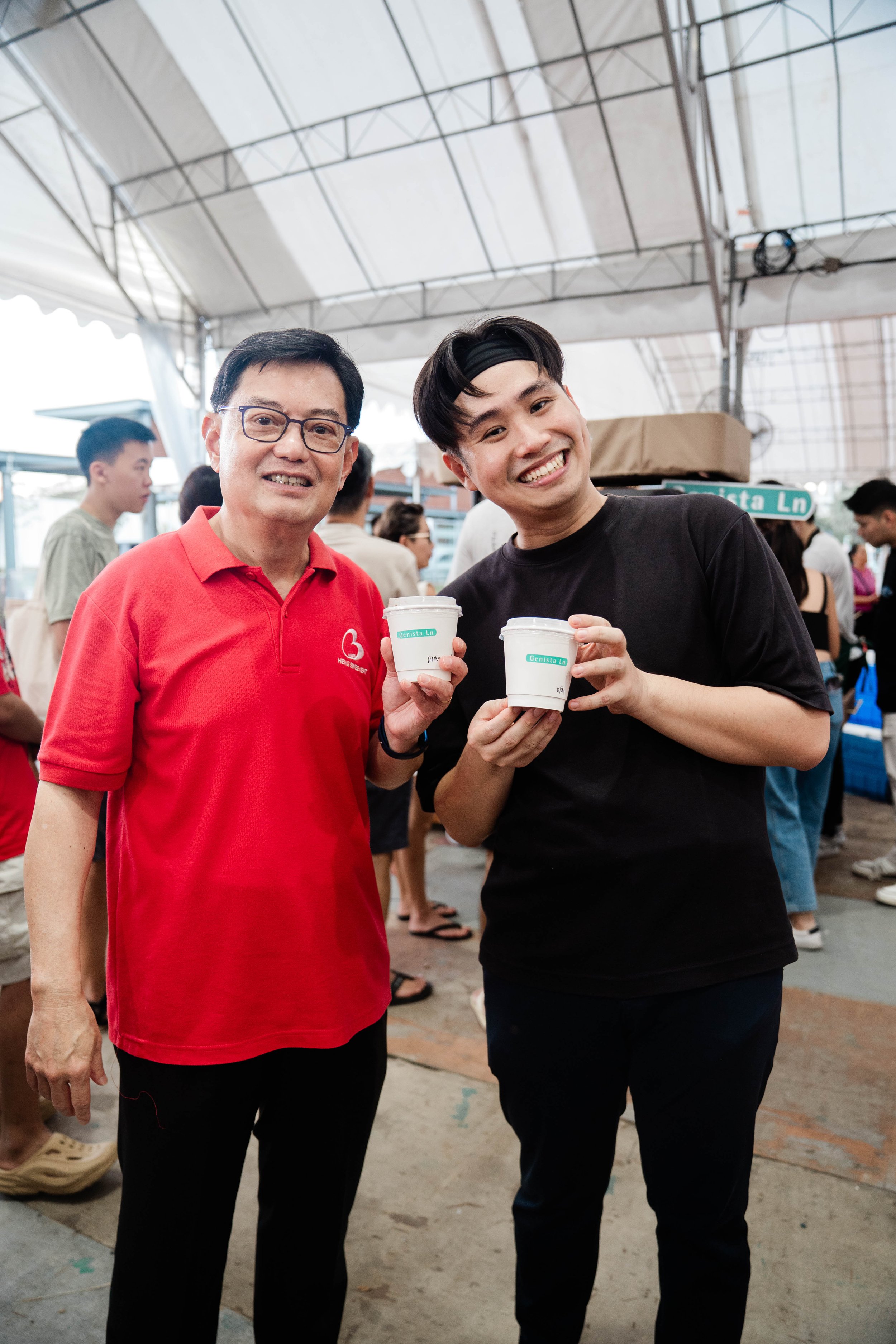 Two men smiling and holding cups of coffee at a busy outdoor gathering under a tent.