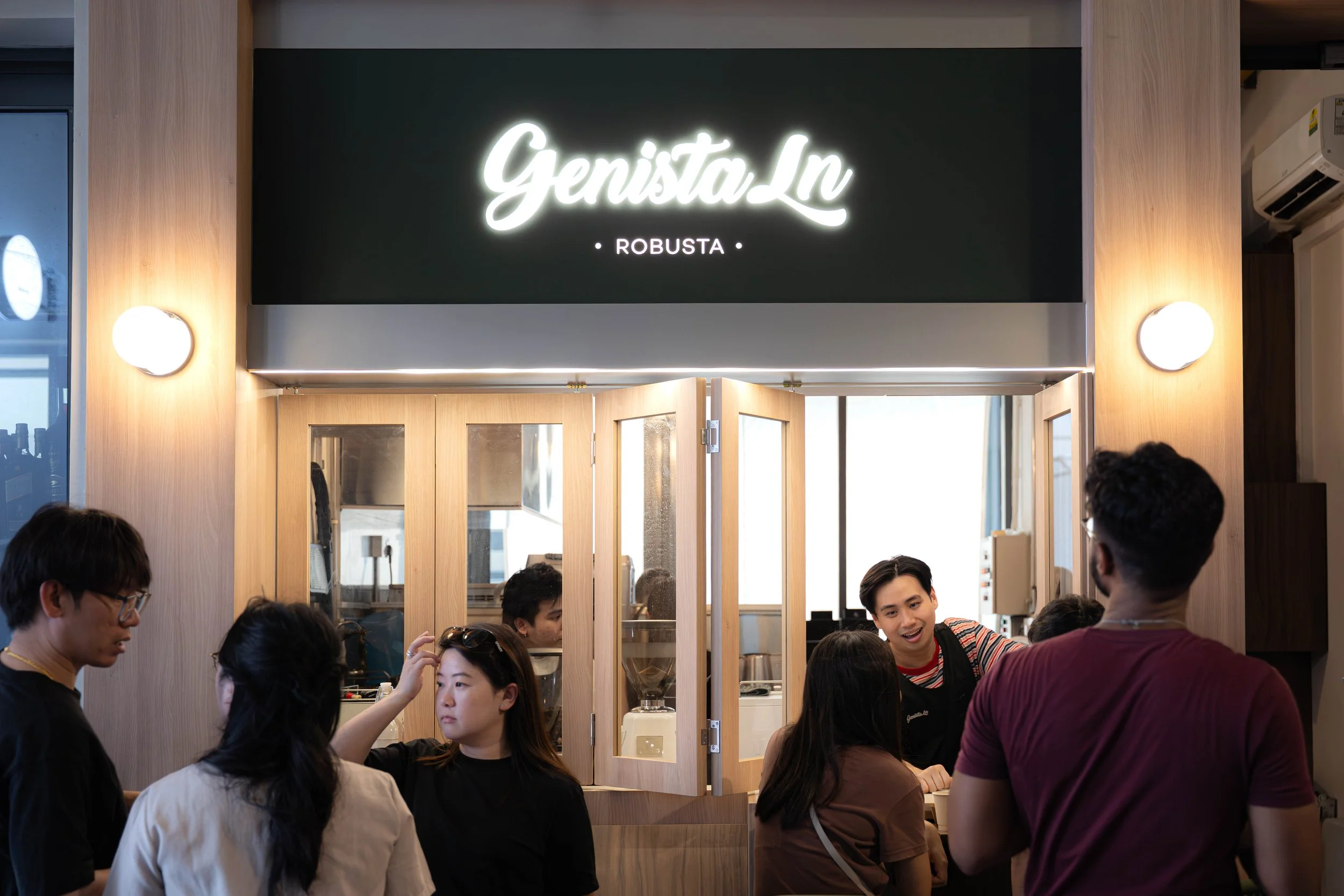 People inside a modern coffee shop called 'Genista Ln Robusta', engaging with baristas, with a large illuminated sign above the counter, wooden accents, and backlit windows.