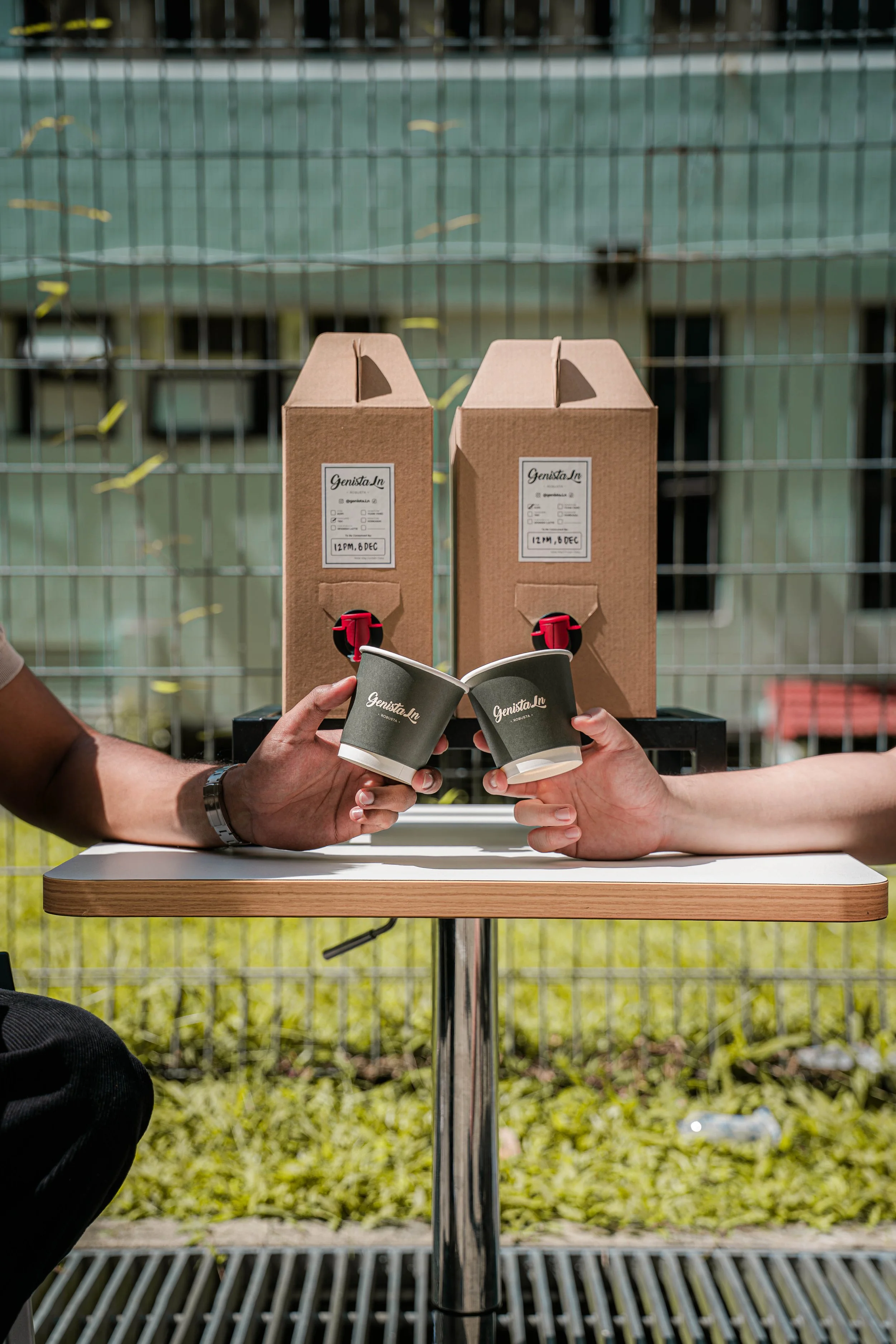 Two people clinking coffee cups, with coffee dispensers in the background at an outdoor setting.