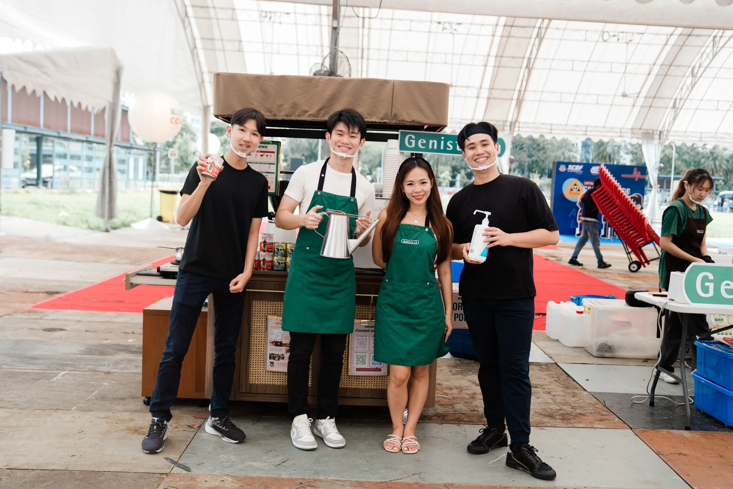 Five people standing in front of a food stall at an outdoor event, smiling and holding various items. The stall has a sign that says "Geniso."