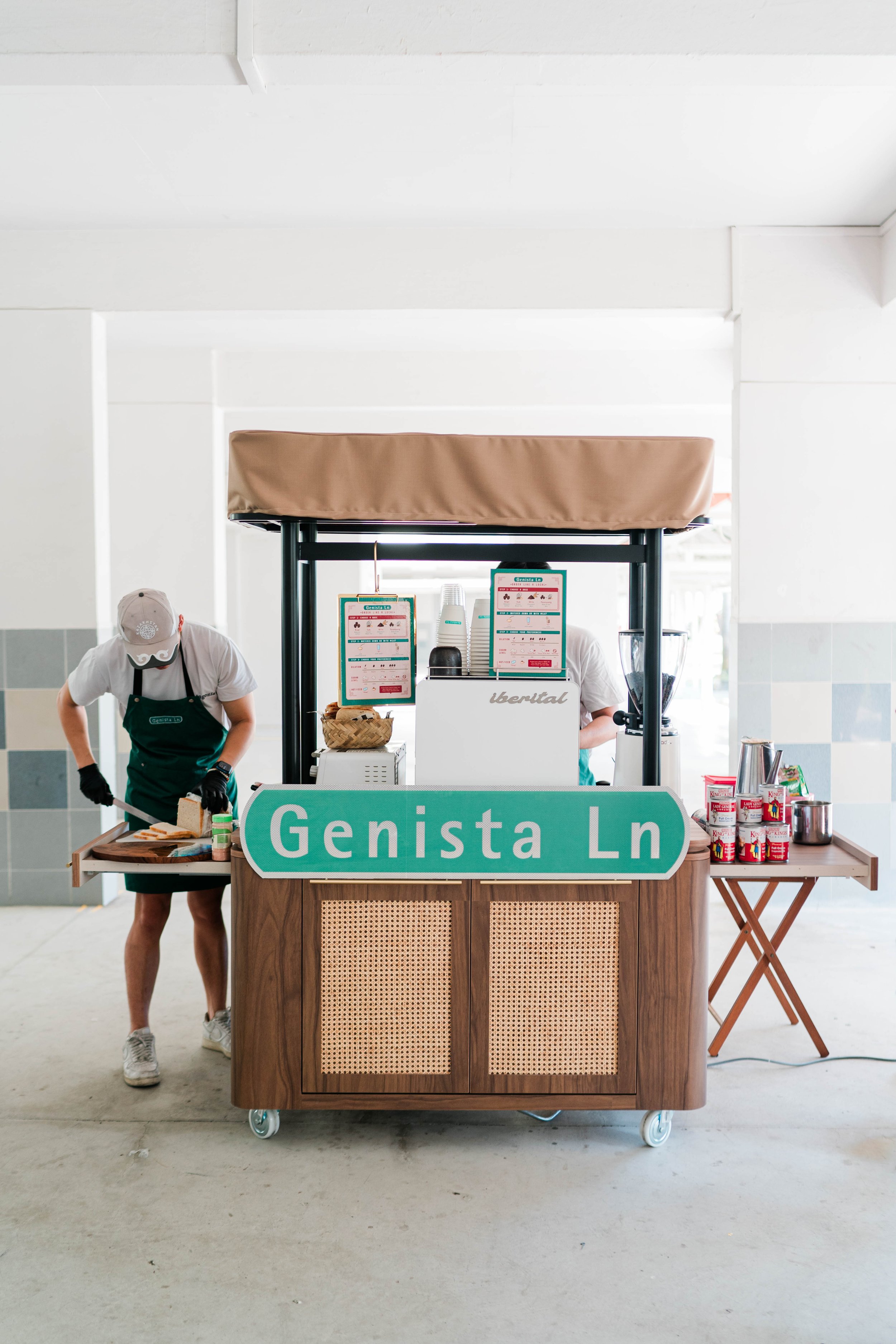 Kopi cart with the sign 'Genista Ln', two workers preparing sandwiches, cups, and canned drinks on the cart.