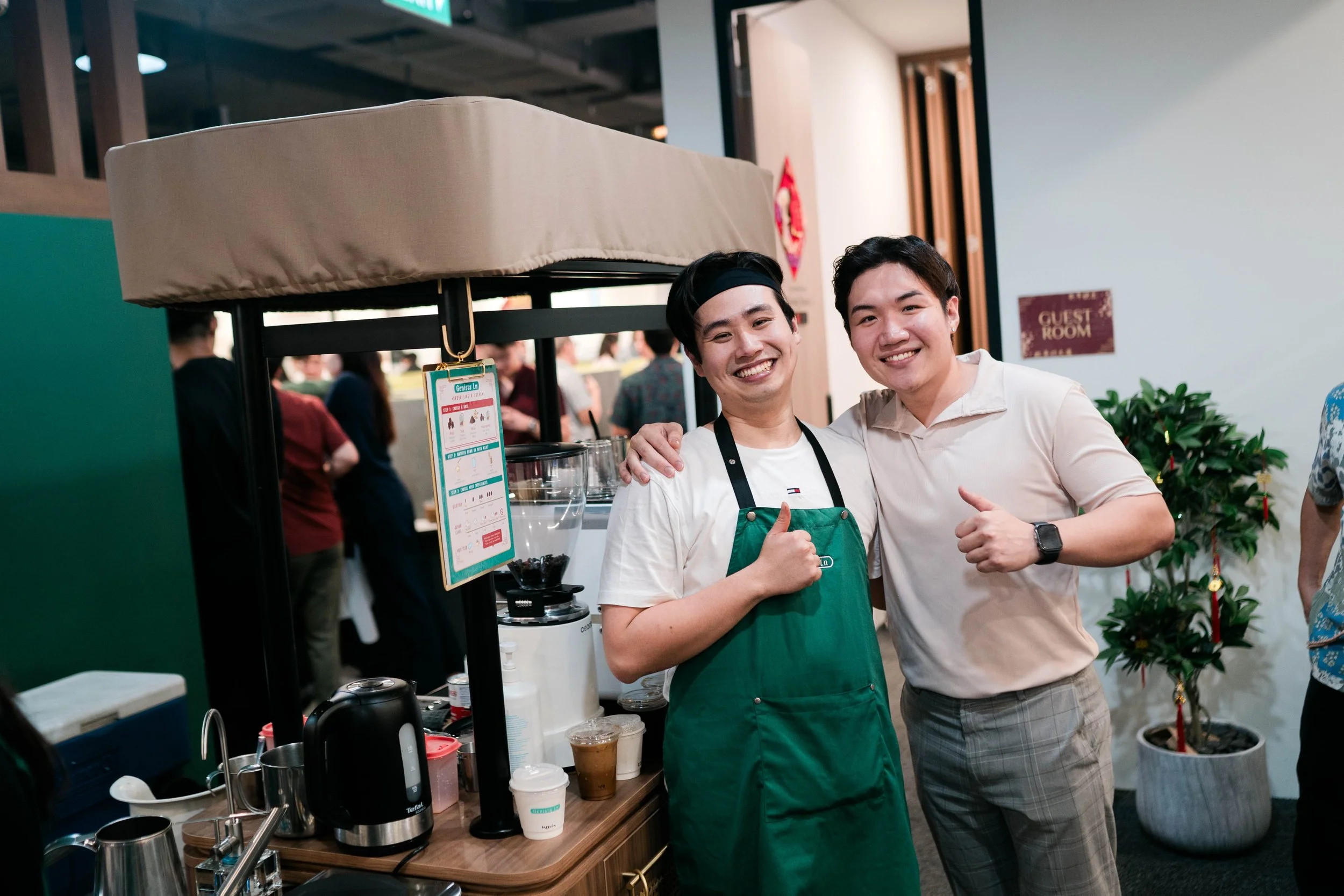 Two men smiling and giving thumbs-up while standing inside a coffee shop. One wears a green apron and black headband, the other a beige polo shirt. Behind them are coffee-making equipment and a potted plant.