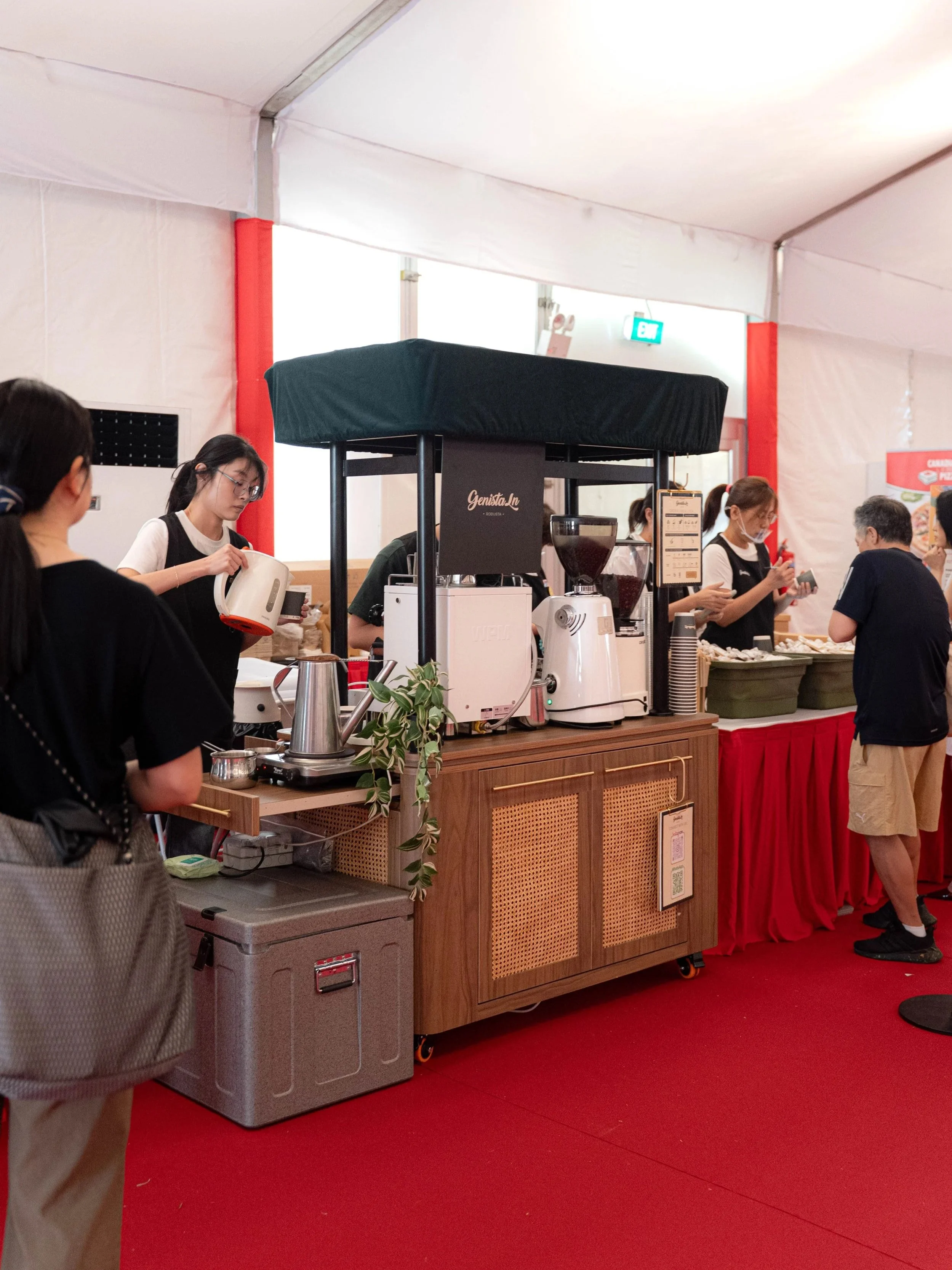 Genista Ln Kopi Cart at a busy Singapore Istana community event with baristas and customers, red and white decor, and coffee equipment.