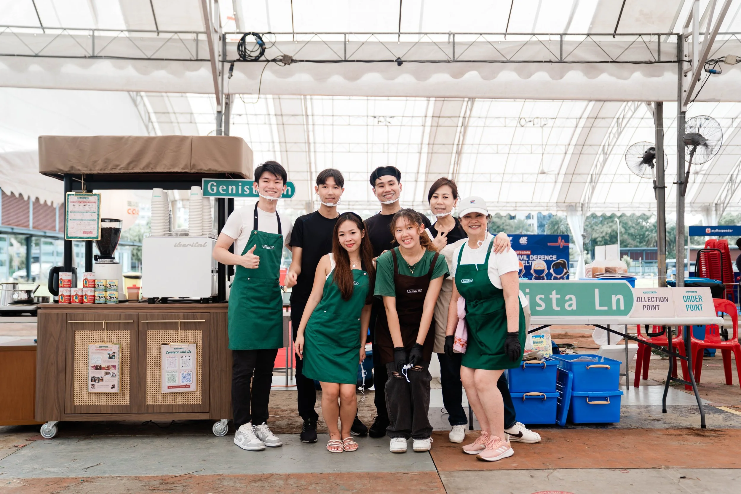 Group of seven people, some wearing green aprons, standing in front of a coffee stand inside a large tent. They are smiling and posing for the camera, with a sign that says "Genista Ln" nearby.