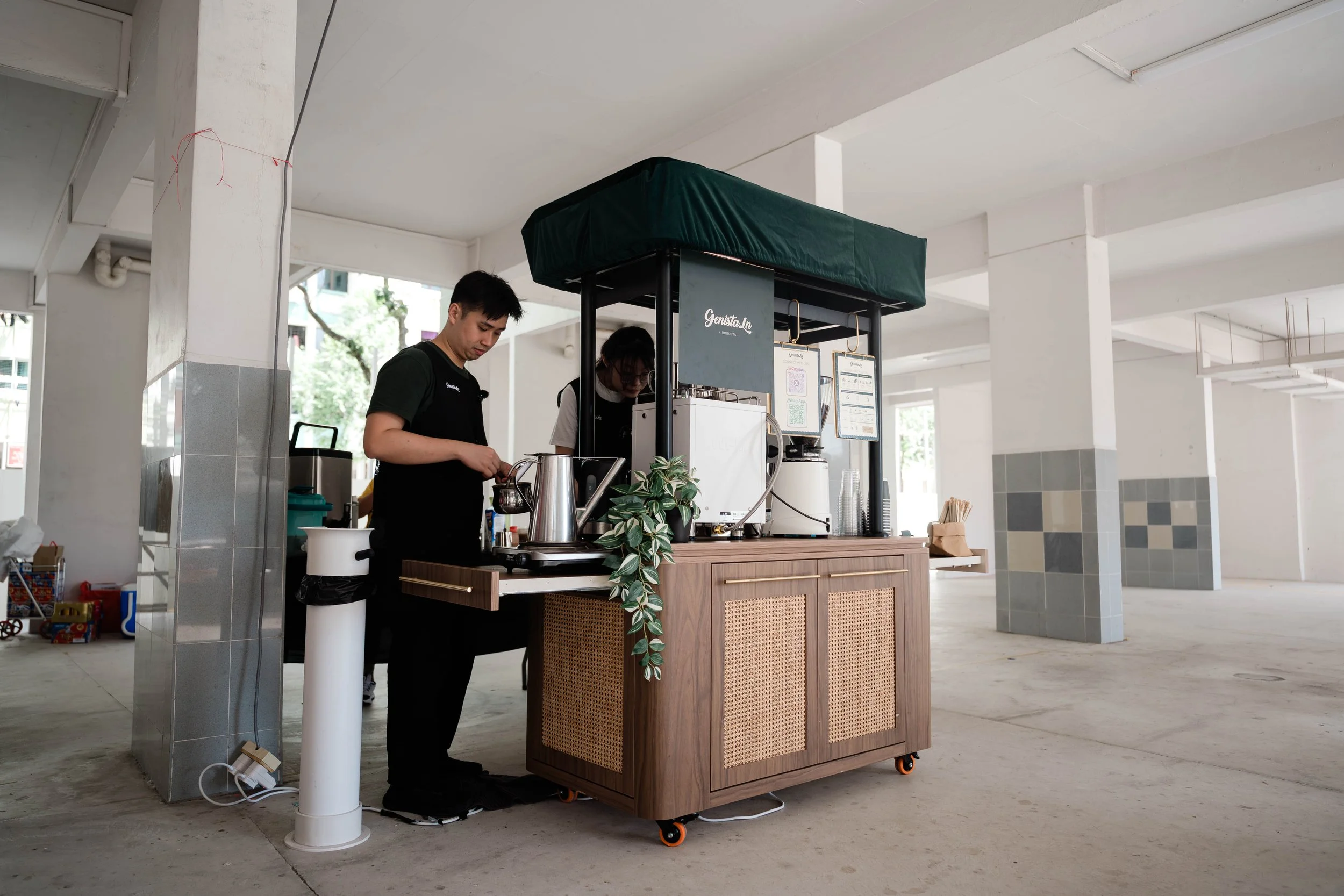 A small coffee stand set up in an open, airy space with two staff members preparing beverages. The stand has a wooden base, a black canopy, and menus hanging on the side.