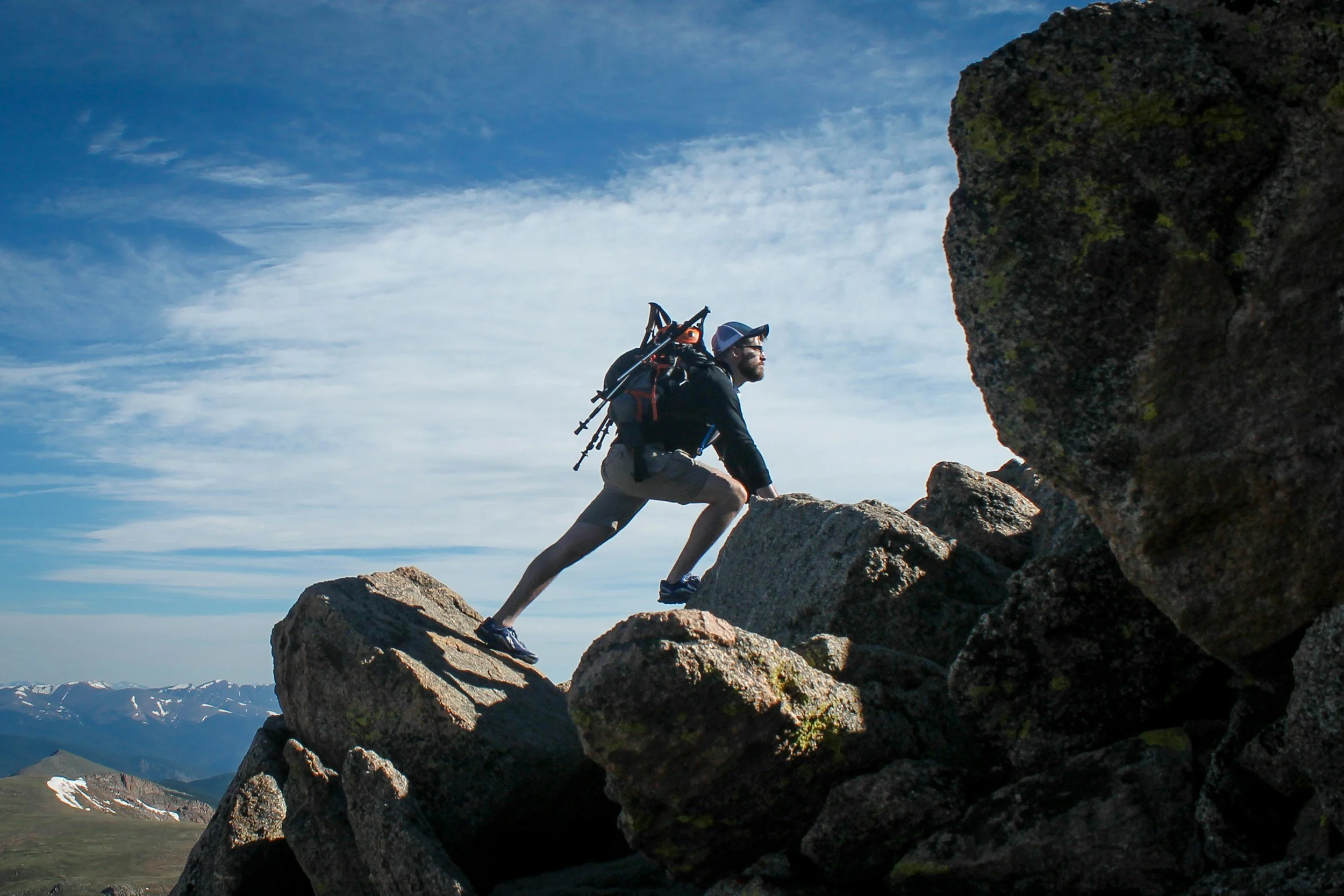 A man climbing rocks outdoors with a backpack and hiking gear under a partly cloudy sky.