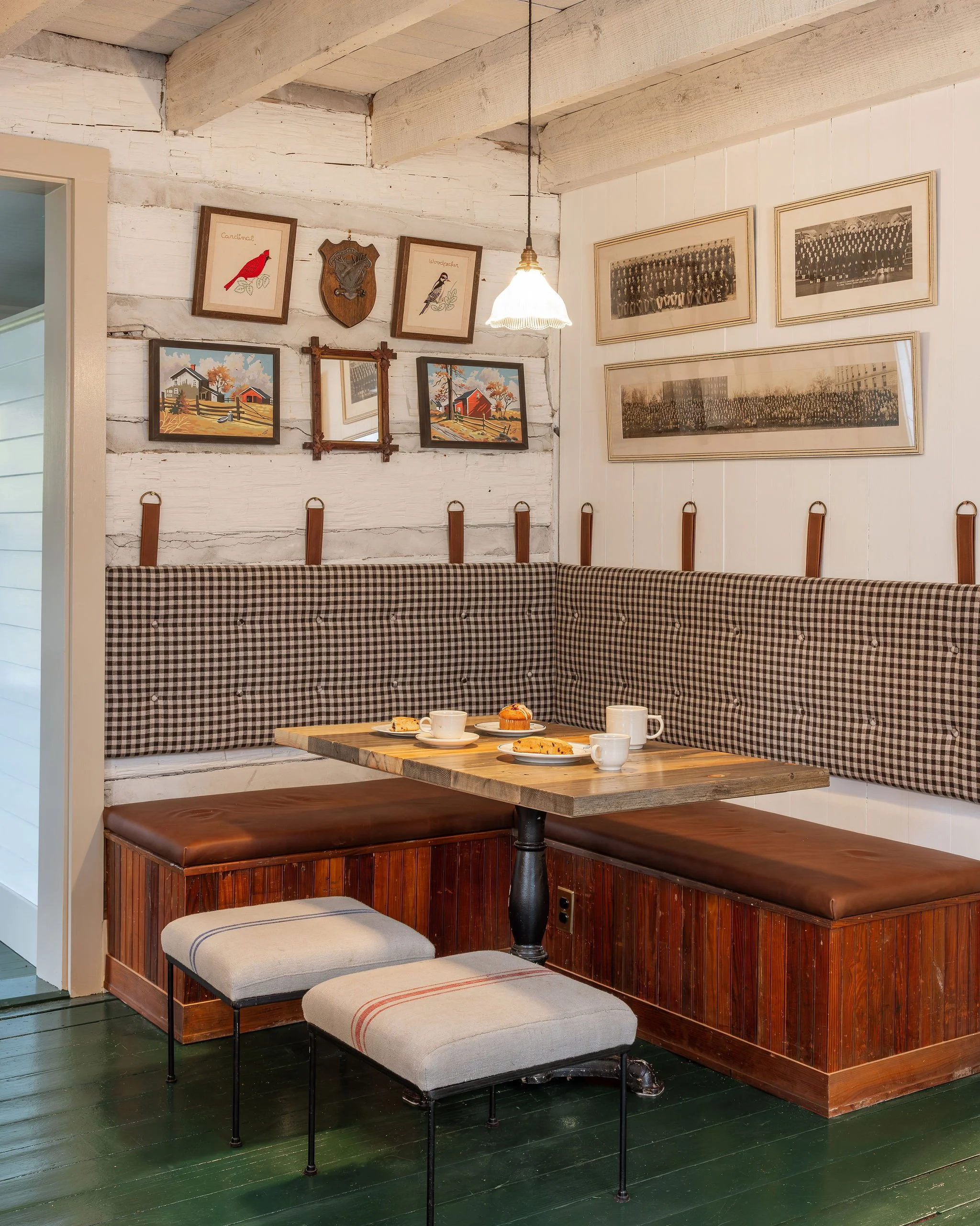 Cozy corner of a cafe with a wooden table, two upholstered stools, and a bench with a checked cushion. The wall behind features framed vintage photos and artwork, with a hanging light fixture overhead.