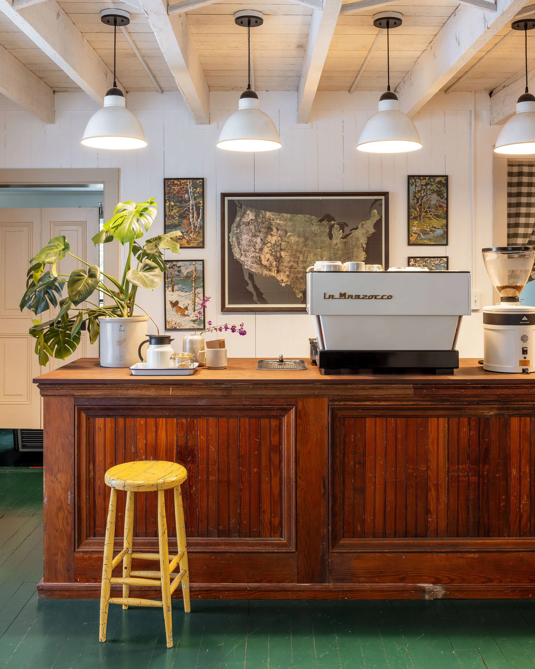 A wooden cafe counter with coffee machine, grinder, and potted plant, decorated with framed artwork and map on white paneled wall, illuminated by hanging lights.